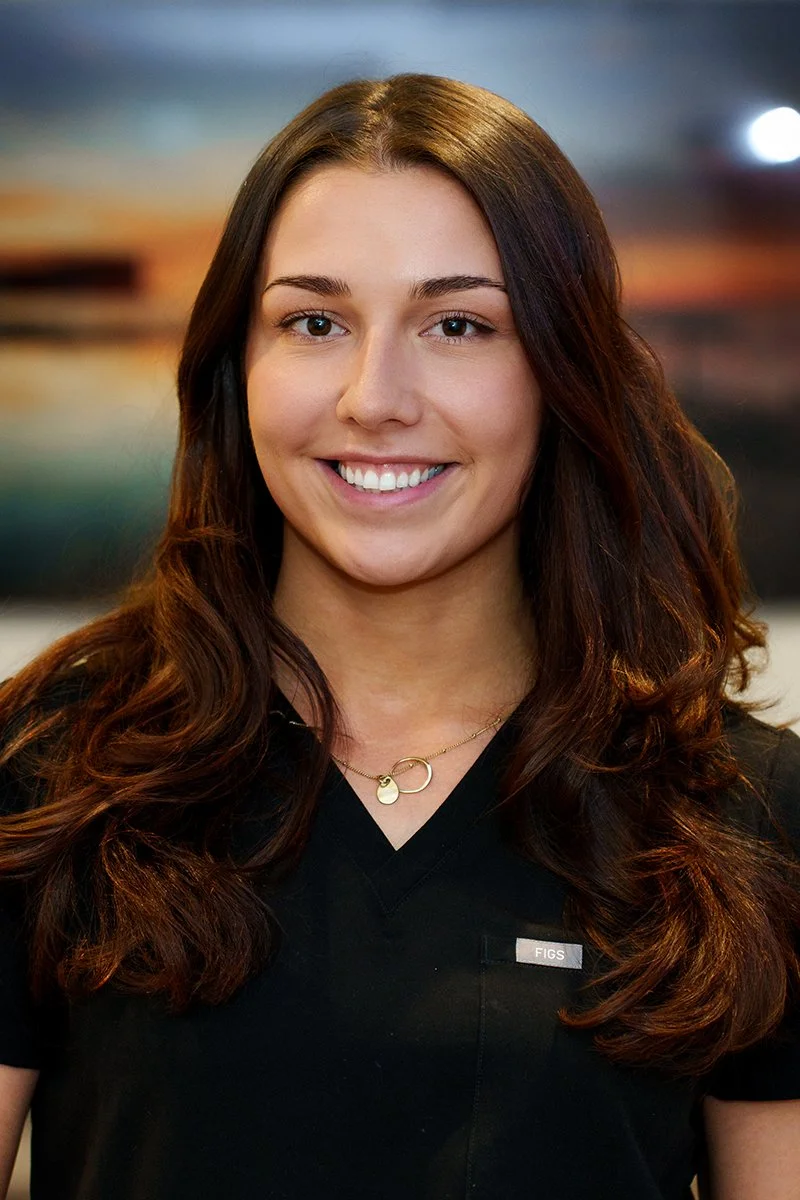 A young woman with long brown hair and a black shirt smiling in front of a blurred sunset background.