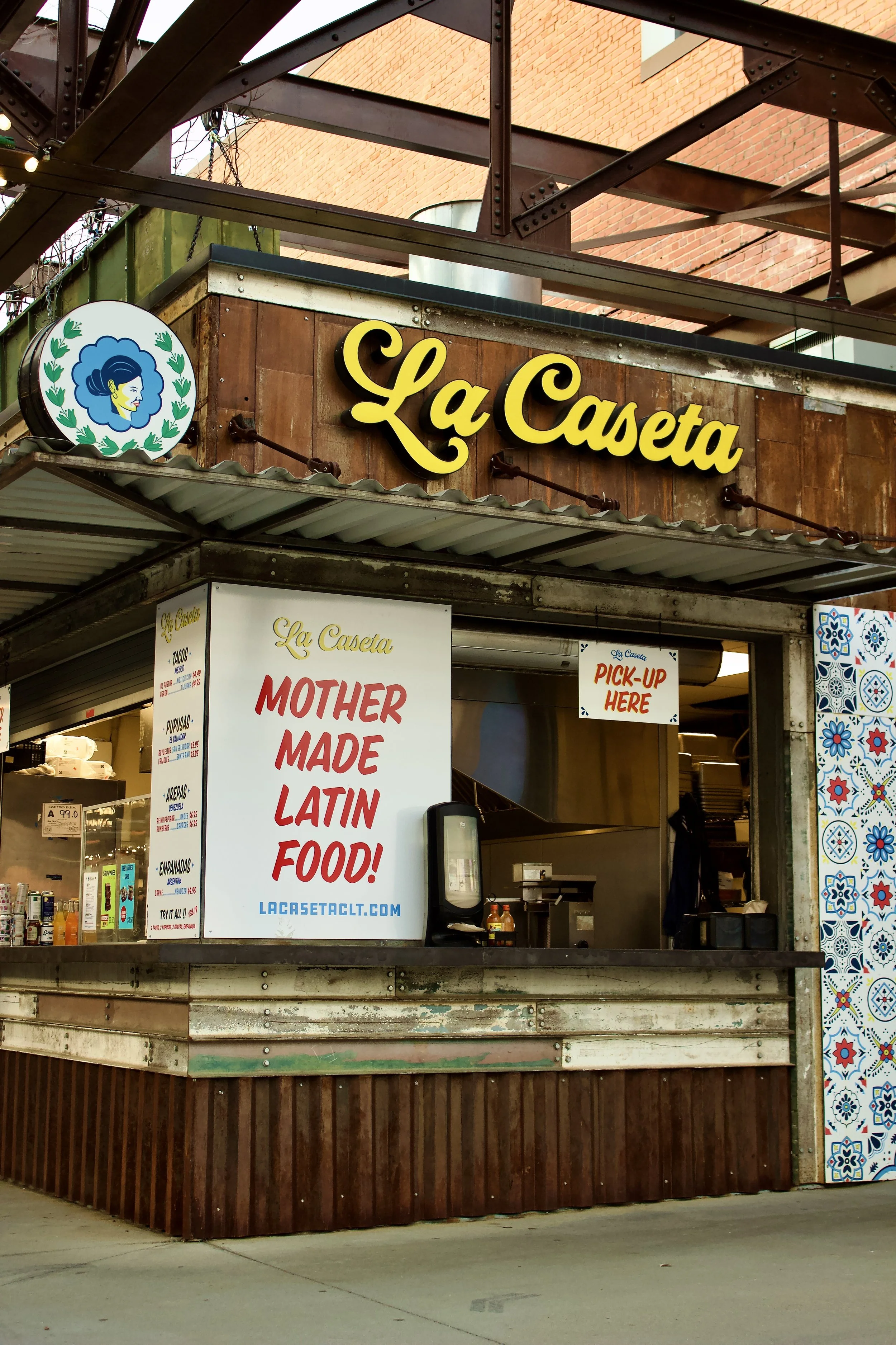 The photo shows a food stand named La Caseta with a sign that advertises Mexican food. The stand has a rustic wooden exterior with a corrugated metal roof and a digital menu display. There is a small window for ordering with a sign that says "PICK-UP HERE". The background includes a brick building and colorful decorative tiles on the side of the stand.