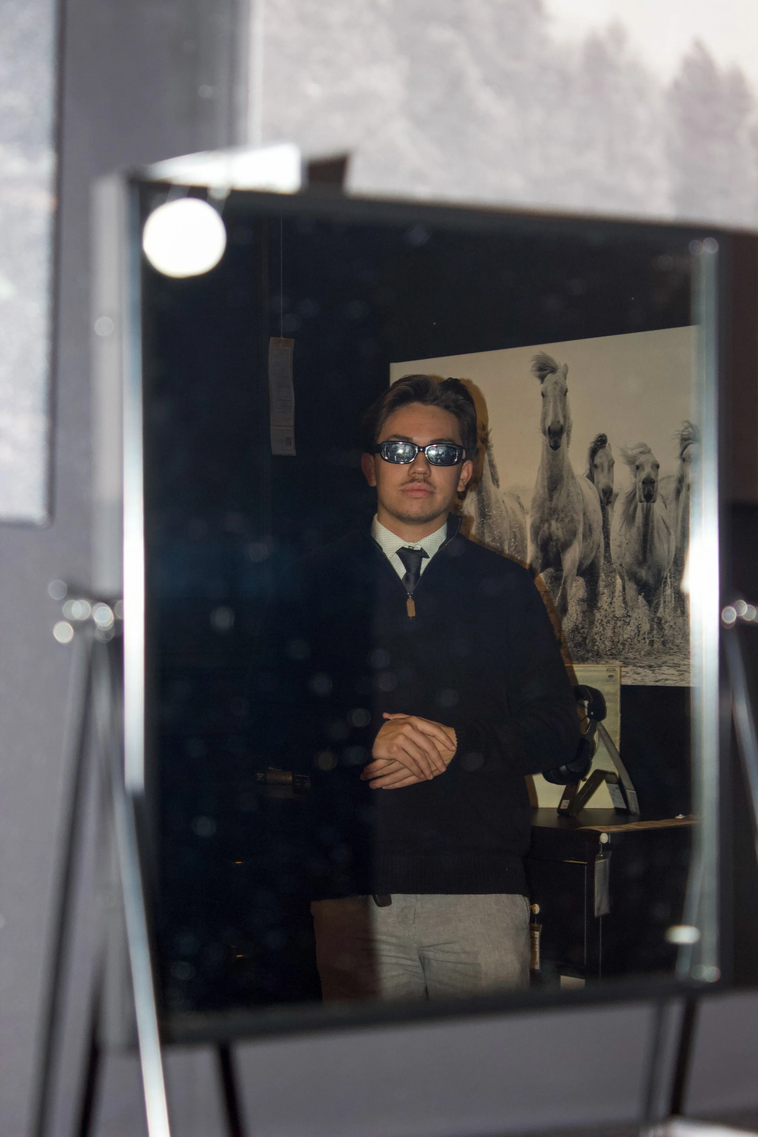 A man wearing sunglasses and a black jacket stands with his hands folded over a counter, reflected in a mirror. Behind him, there is a bedroom or office with a black and white horse-themed poster on the wall.