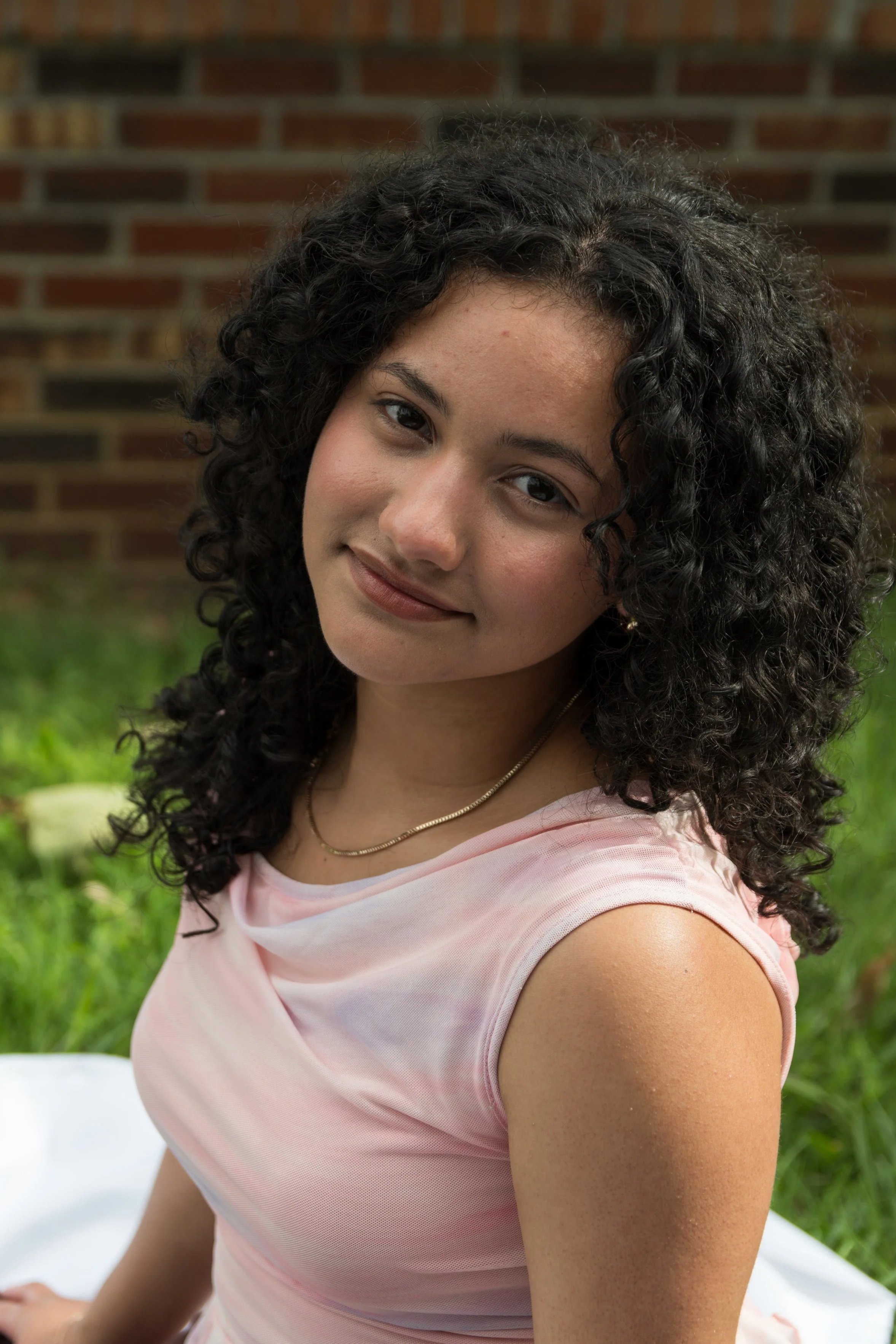 A young woman with curly dark hair smiling outdoors in front of a brick wall and greenery, wearing a light pink top and a necklace.