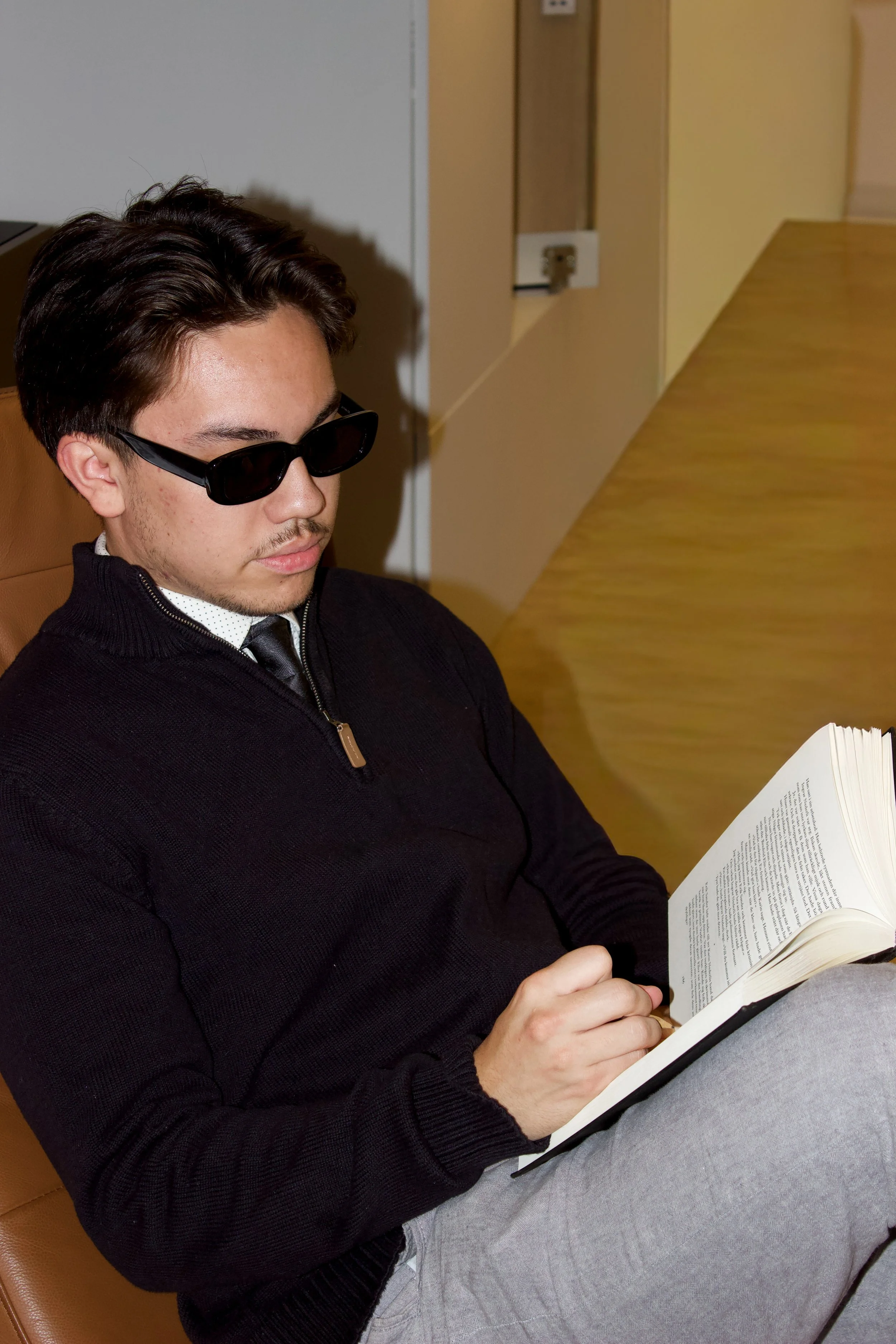 A young man with dark hair, wearing sunglasses, a black sweater, a white shirt with black dots, a tie, and gray pants, sitting in a beige chair in a room with yellow walls, reading a book.
