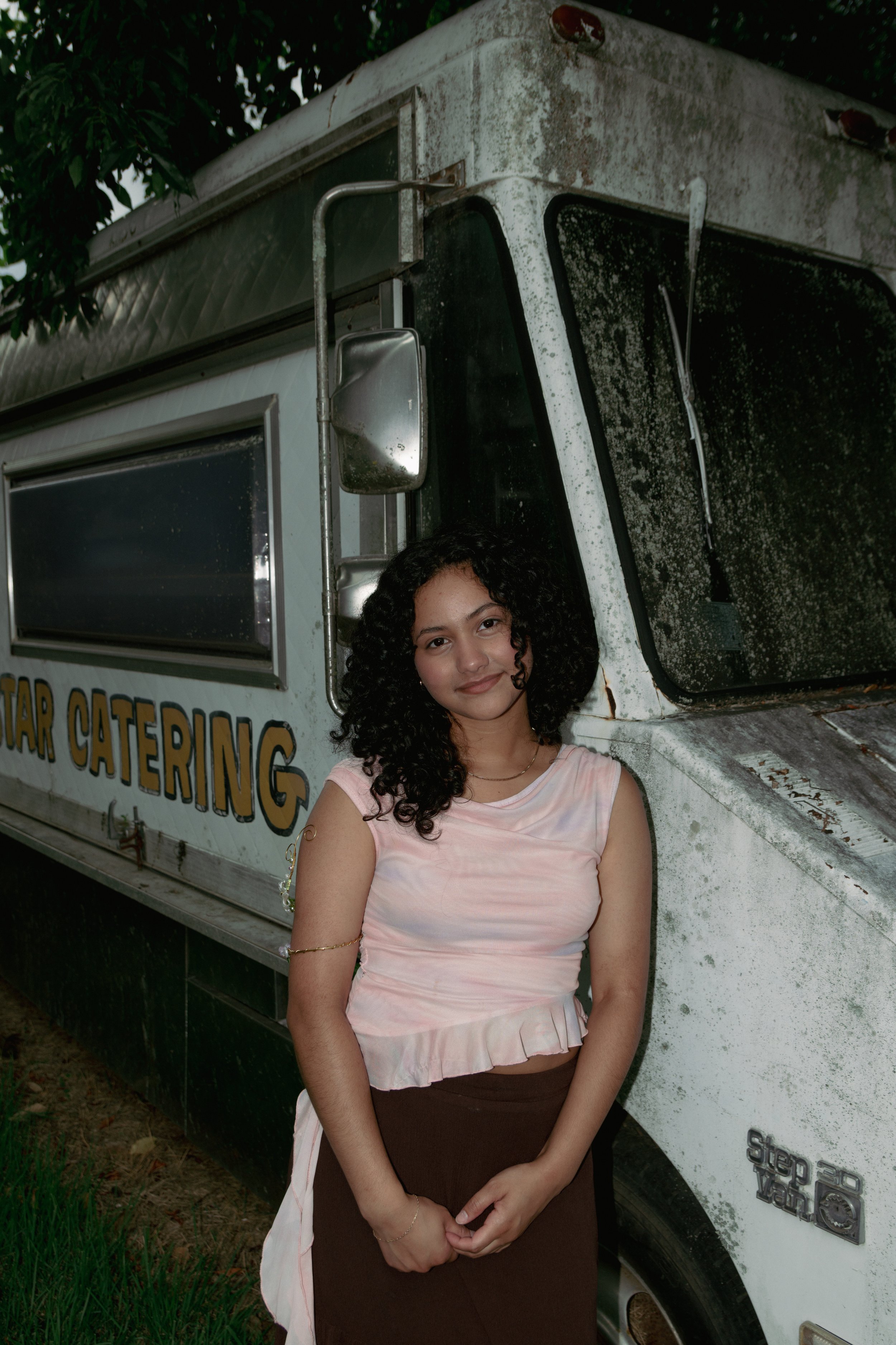 A young woman with curly dark hair standing in front of a weathered food truck. She is wearing a light pink top and a brown skirt, with an off-white cloth draped over her arm. The food truck has a window and a sign partial reading 'STAR CATERING,' and appears old and dirty with dirt and grime on its surface.