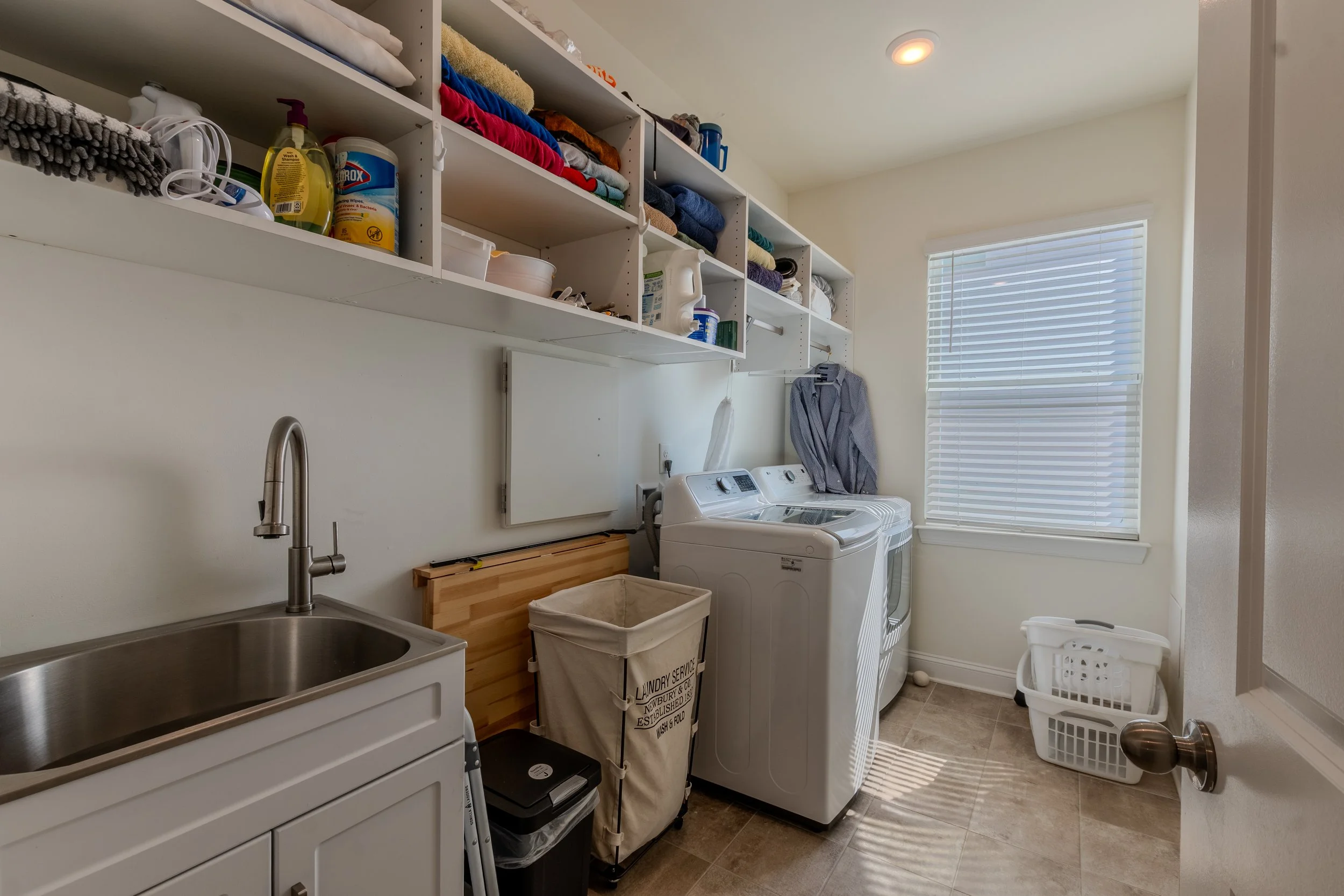 A laundry room with shelves filled with folded towels and laundry supplies. There are washing machine and dryer units, laundry baskets, a sink, and a window with blinds.