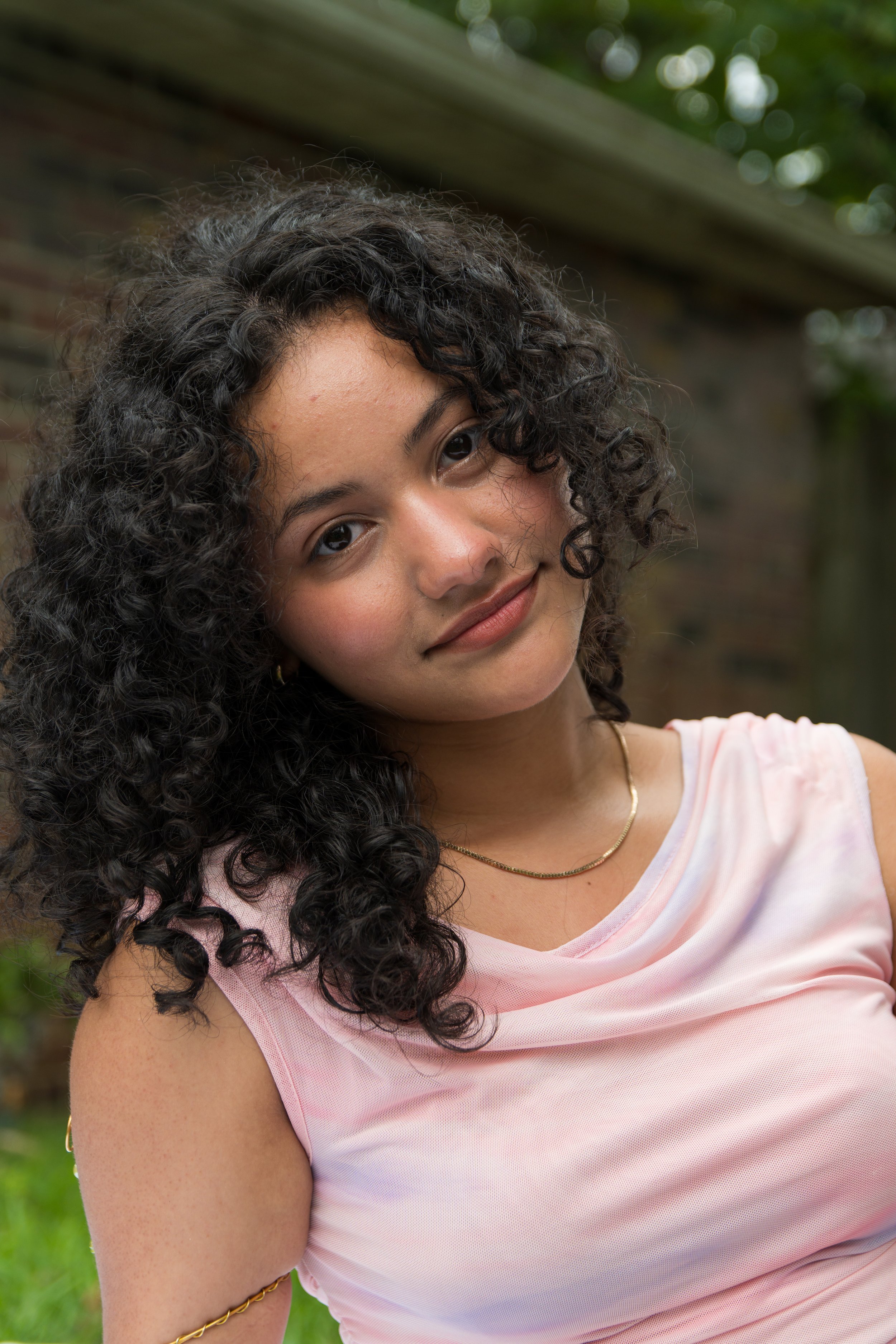 A woman with dark curly hair, wearing a pink top, gold necklace, and gold bracelet, is outdoors with a blurred green and brick background.