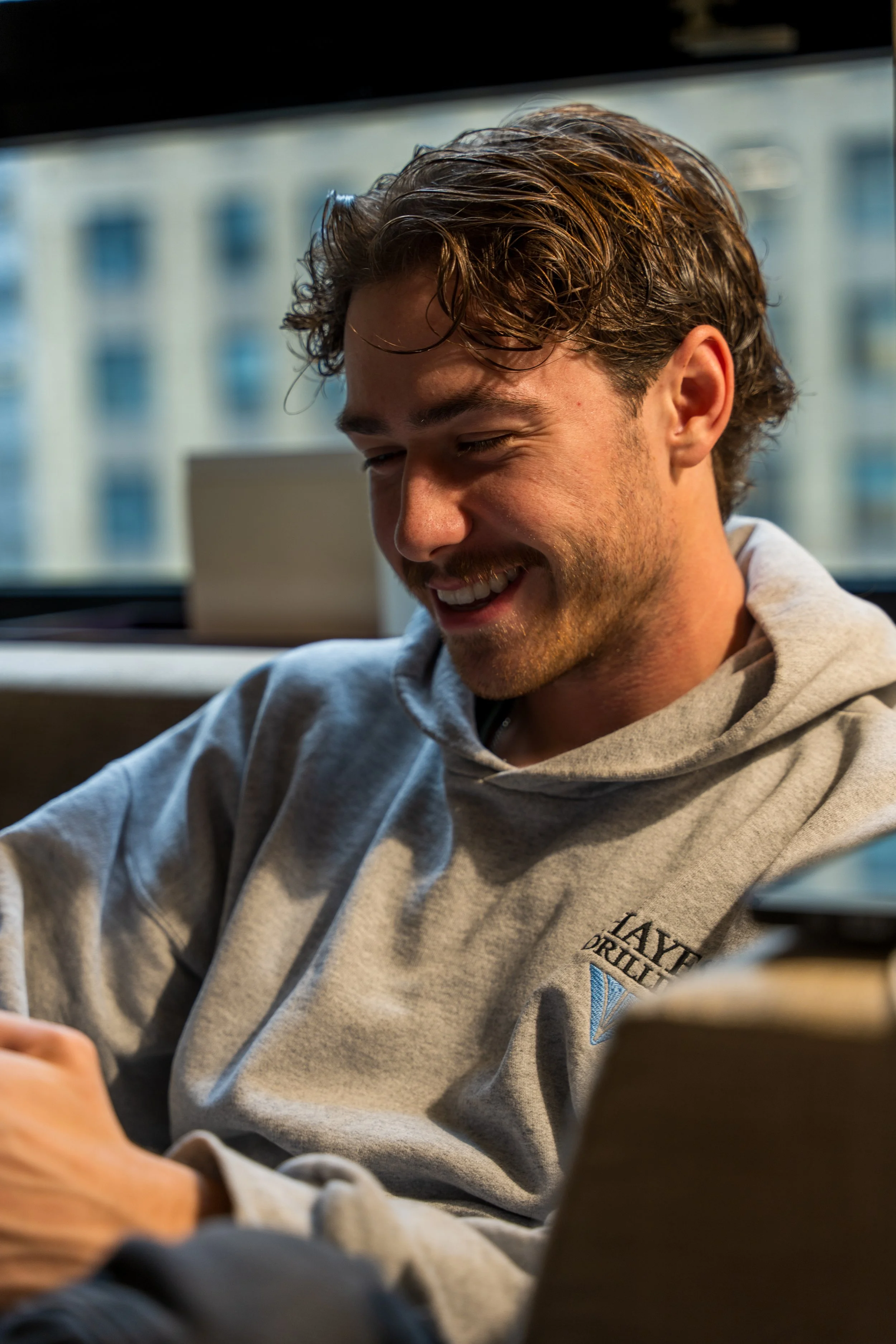 A young man with curly hair smiling, wearing a gray hoodie, sitting indoors with a blurred cityscape window behind him.