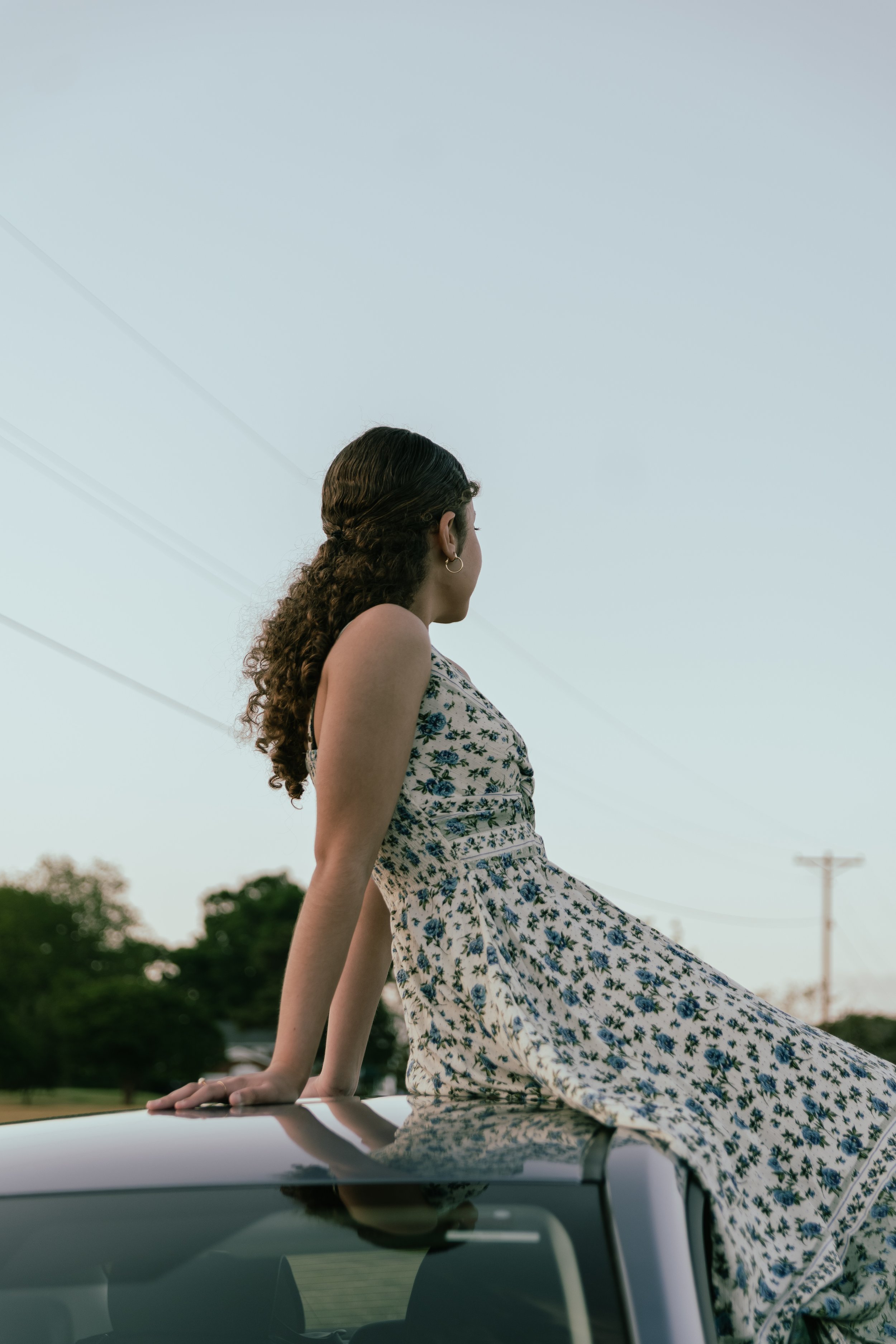 A young woman with curly hair wearing a floral dress is sitting on the roof of a car, looking away from the camera, with a background of trees and power lines, during daytime.