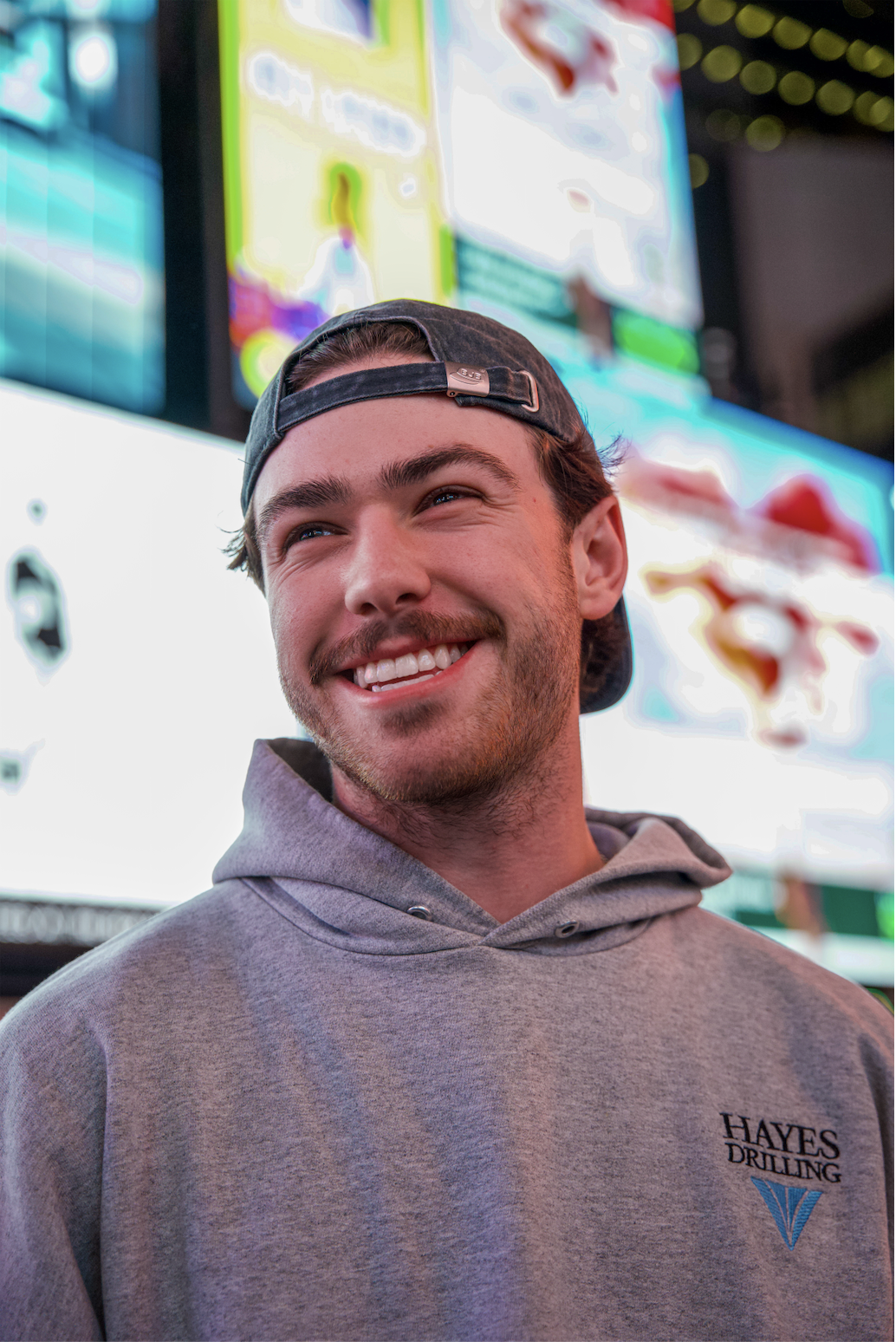 A smiling young man wearing a backwards baseball cap and a gray hoodie with a "Hayes Drilling" logo, standing in front of brightly lit digital billboards at night.