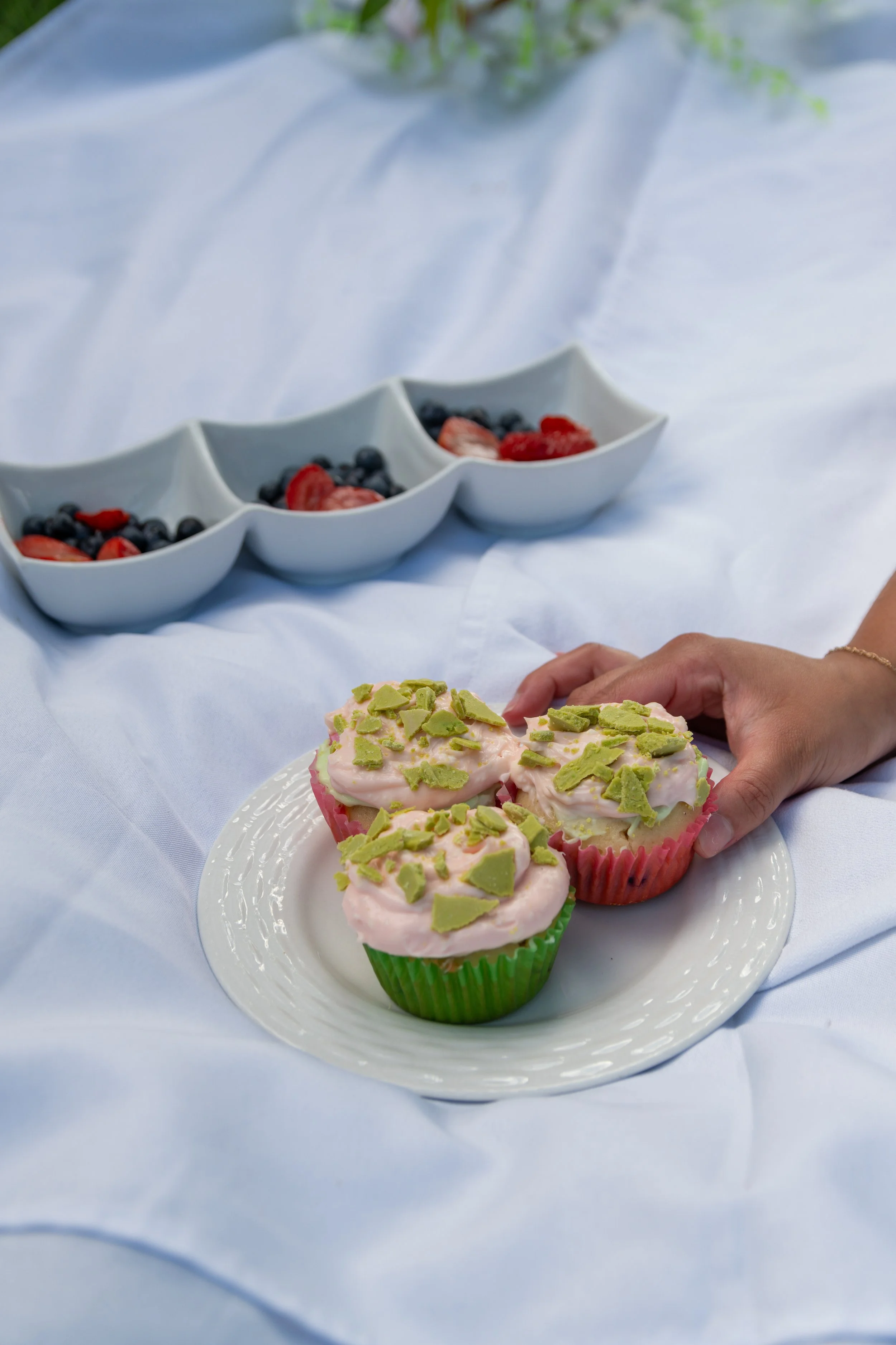 Cupcakes with pink frosting and green sprinkles on a white plate, with a bowls of mixed berries, including strawberries and blueberries, in the background on a white tablecloth.