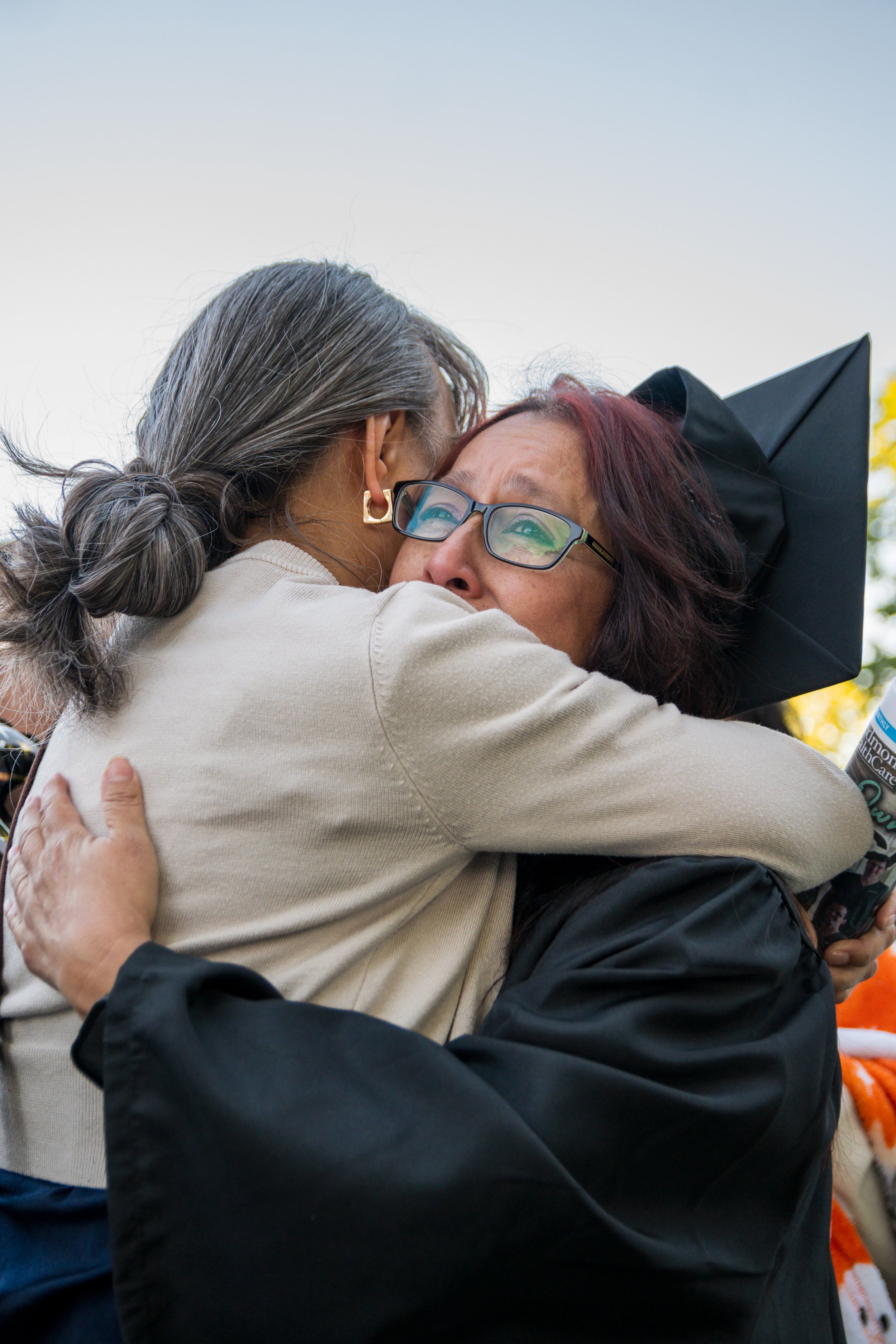 Two women hugging each other, one wearing glasses and a graduation cap, the other with gray hair tied back, outdoors with a clear sky.