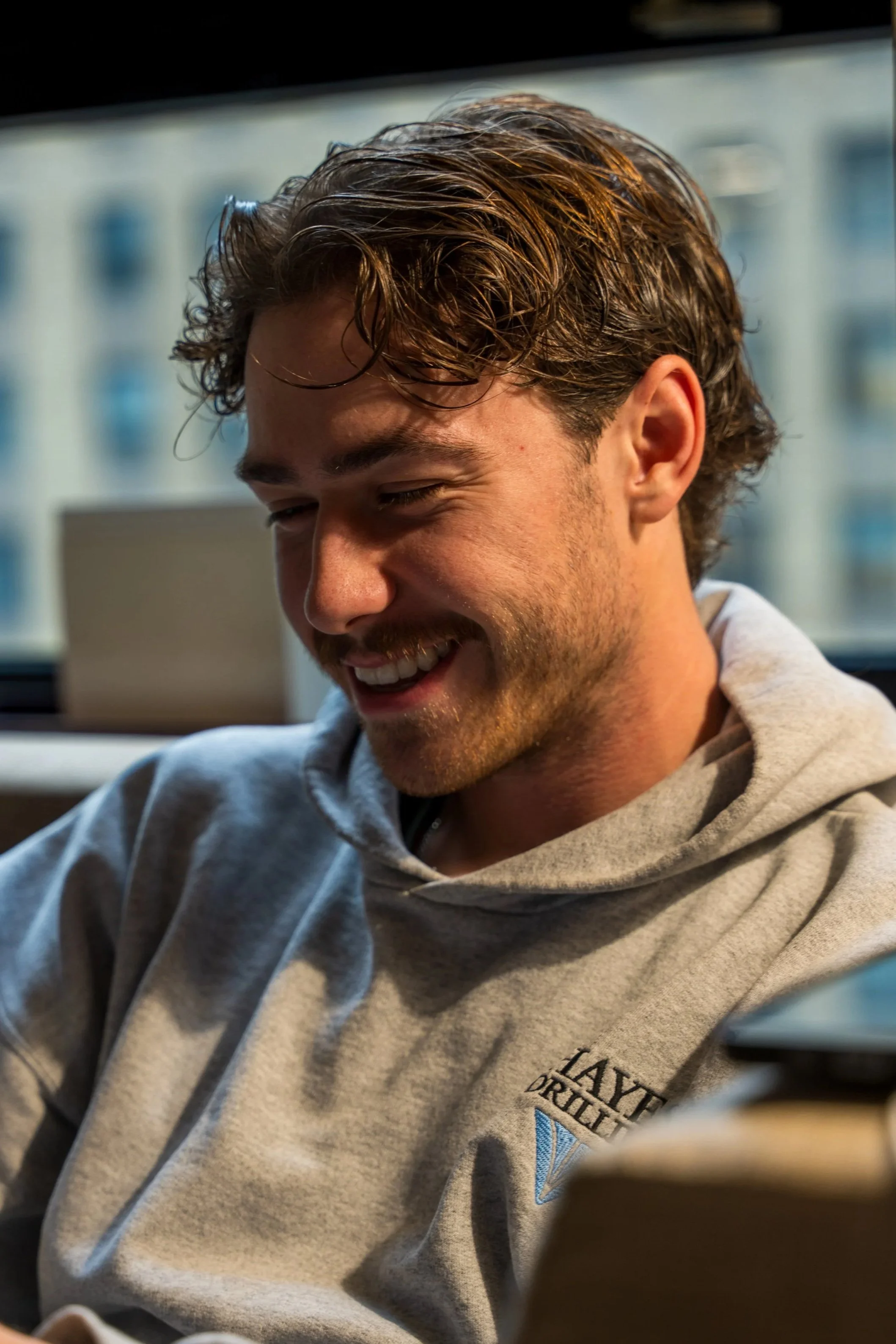 A young man with curly brown hair and a beard, smiling and looking downward, wearing a gray hoodie with a logo on it, sitting indoors with a blurred window background.
