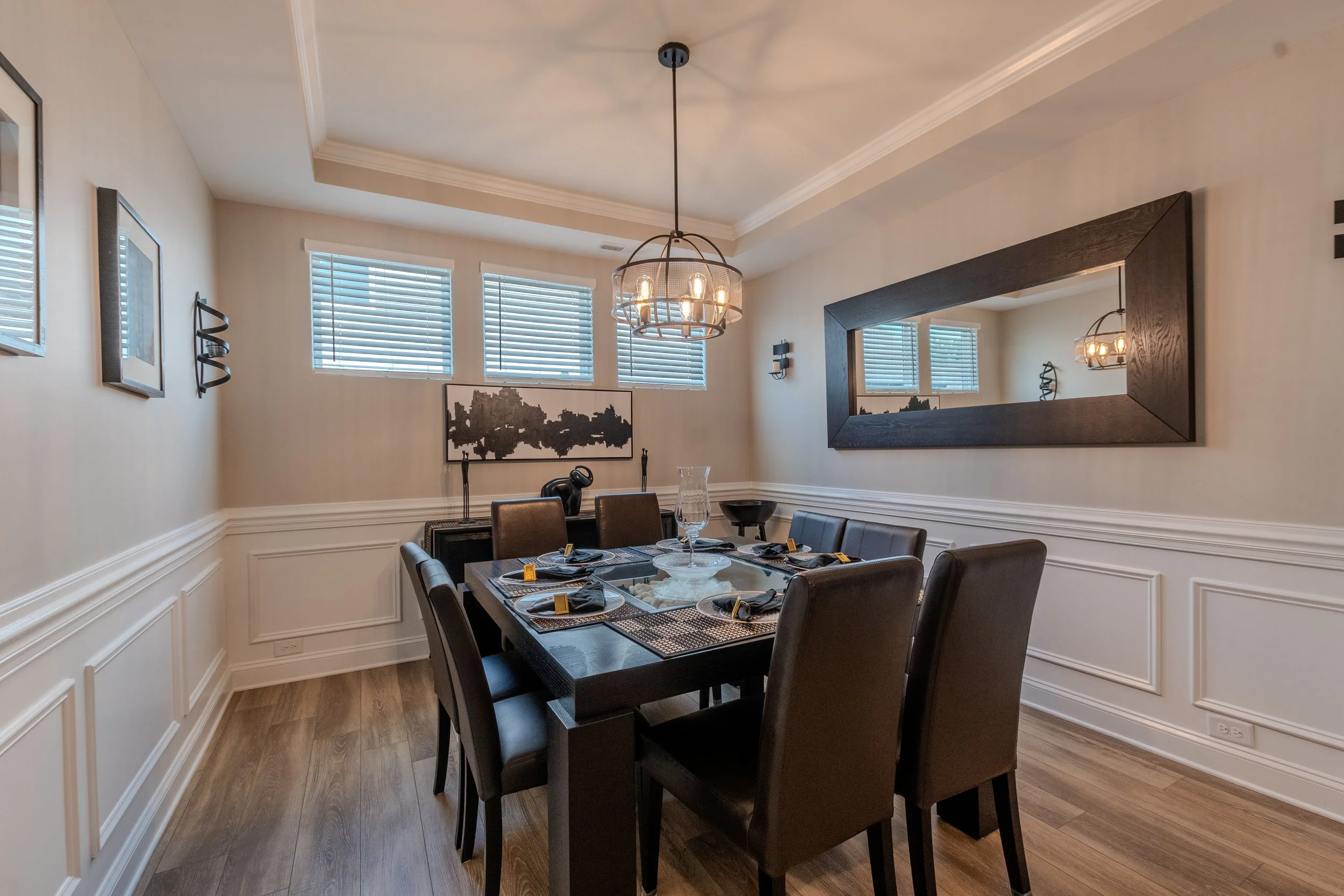 A dining room with wainscoting, a large rectangular mirror with a dark wood frame, a chandelier, and a table set with plates, glasses, and napkins.