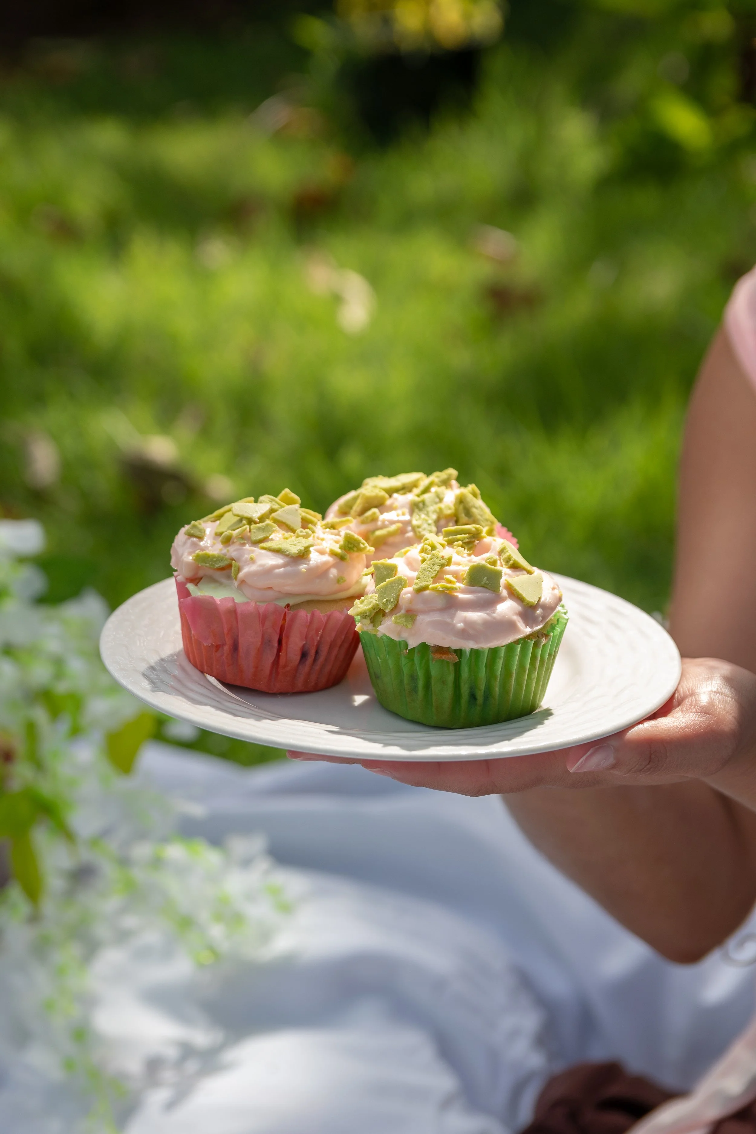 A person holding a white plate with two frosted cupcakes topped with matcha green tea chopped pistachios outdoors with a green blurred background.