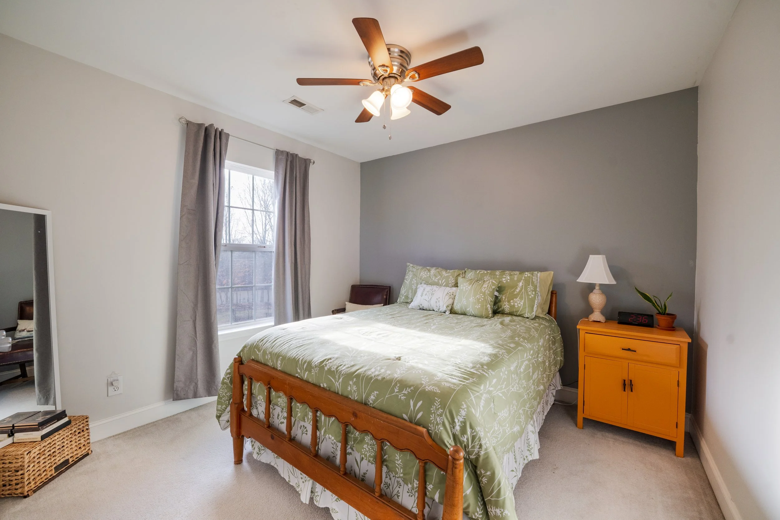 A bedroom with a wooden bed frame, green floral bedding, and matching pillows. There's a yellow nightstand with a lamp and a digital clock. A window with gray curtains is on the wall, and a ceiling fan with wooden blades and lights is above.