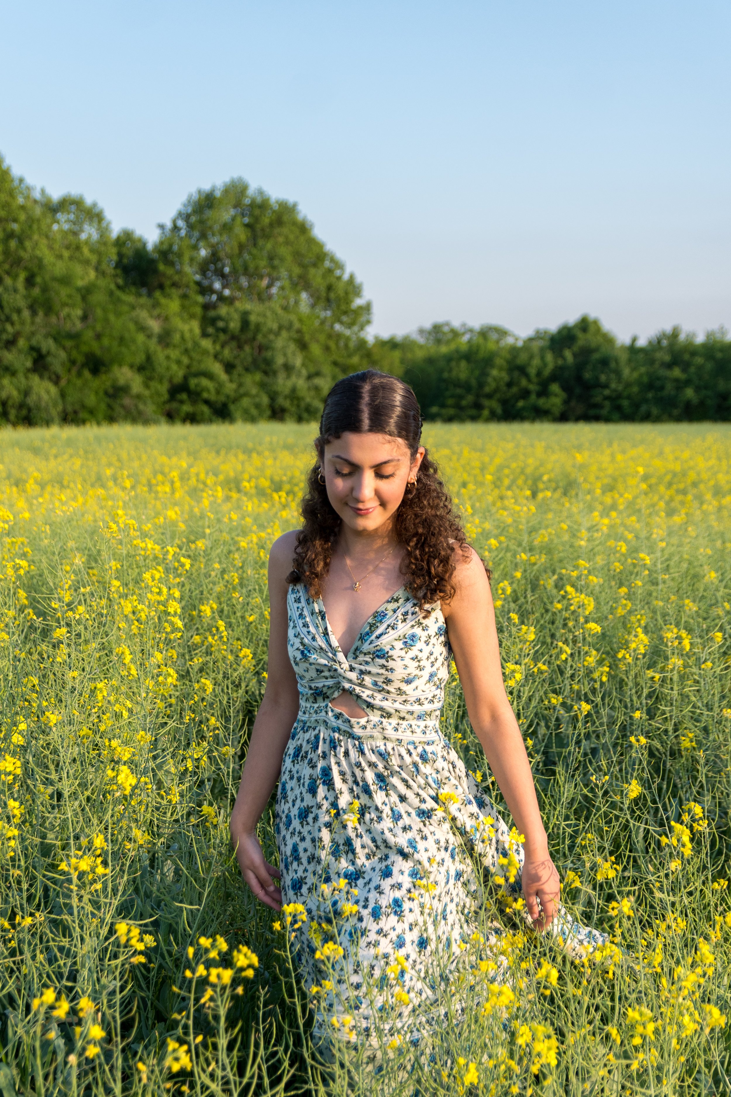 Young woman with curly hair in a floral dress walking through a yellow flower field on a clear day.
