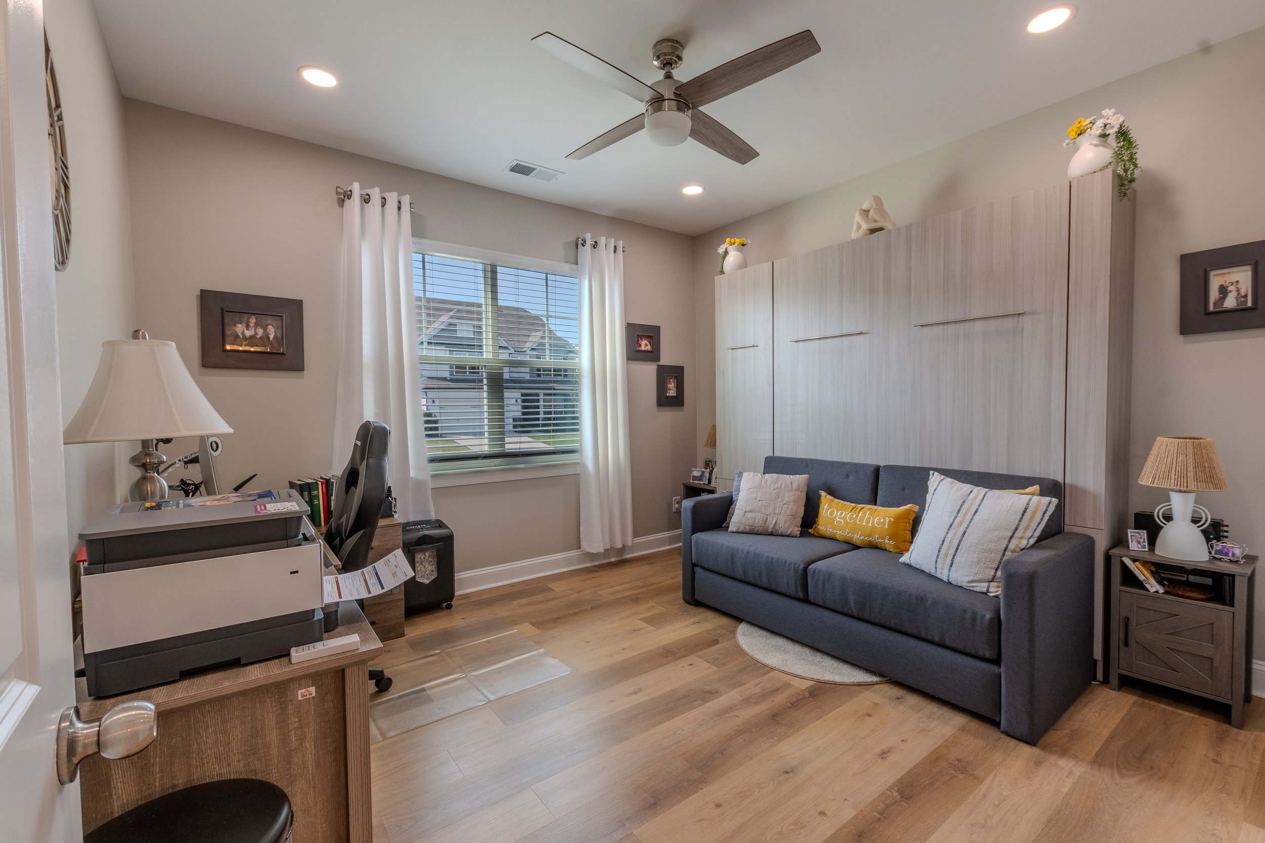 A living room with a gray couch, decorative pillows, a wooden side table, a lamp, a window with white curtains, and a ceiling fan.