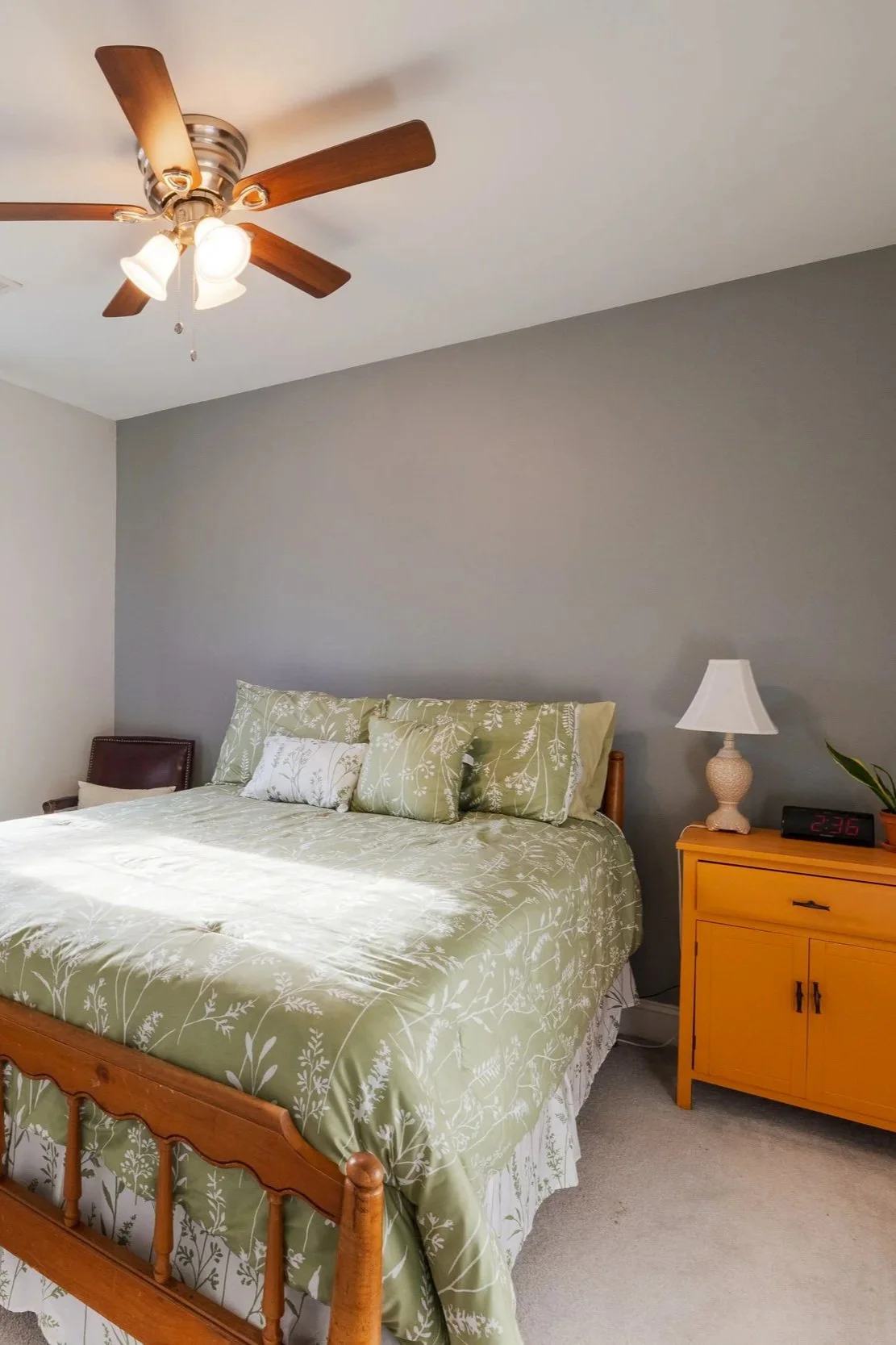 A bedroom with a bed covered in a green, white floral pattern bedspread and pillows, a wooden nightstand with a yellow finish, a white lamp, a digital clock, a plant, and a ceiling fan with wooden blades and three light fixtures.