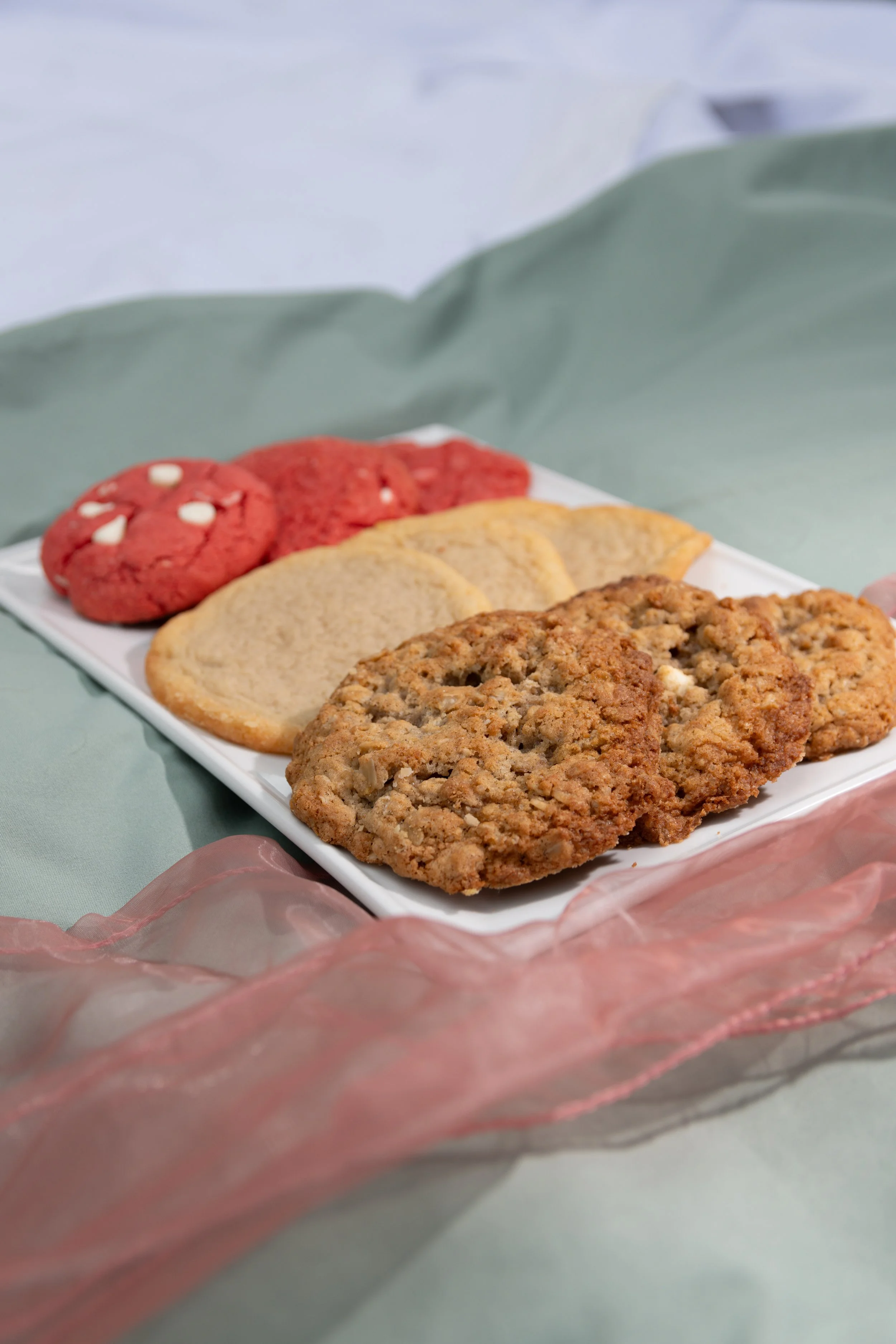 A white rectangular tray with assorted cookies including oatmeal, sugar, and red velvet cookies, held by a person with pink tissue paper around the tray.