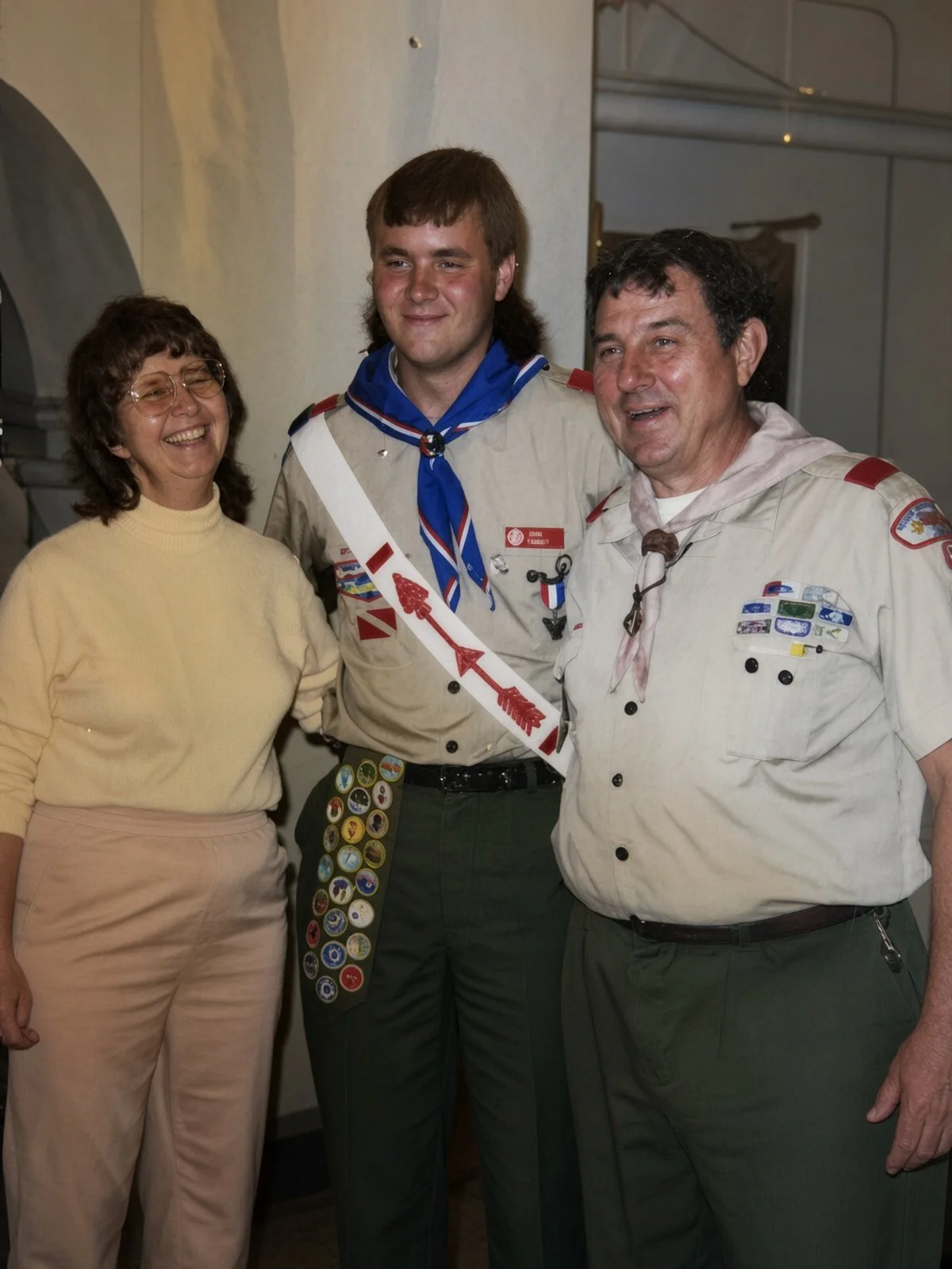 Three people smiling indoors, one woman on the left wearing a yellow turtleneck sweater and light-colored pants, and two men in scout uniforms with various patches and badges, standing close together.