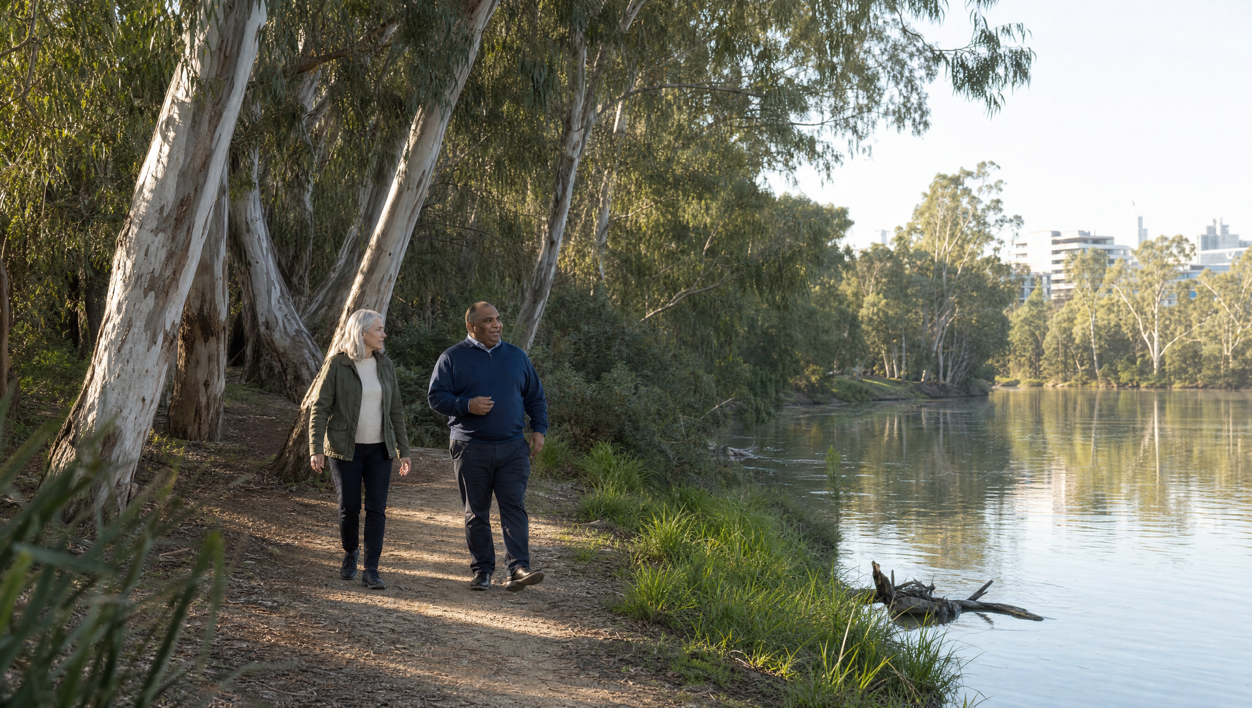 Two people, a woman with gray hair and a man with dark skin, walk along a wooded riverside trail during daytime, surrounded by tall eucalyptus trees and city buildings in the background.
