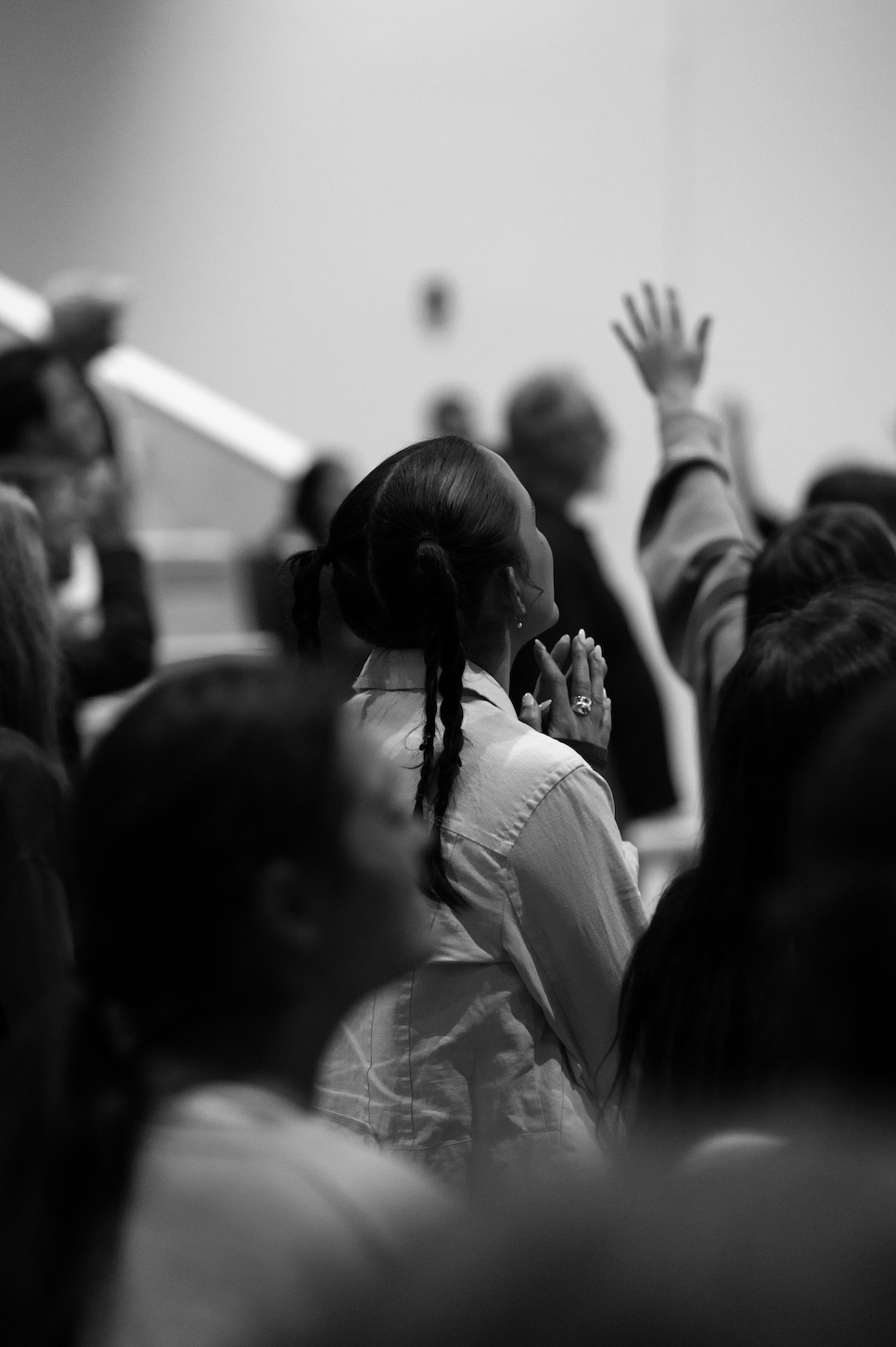 A black and white photo of a group of people attending a church service, with one woman in the center clasping her hands and a person in the background raising their hand.