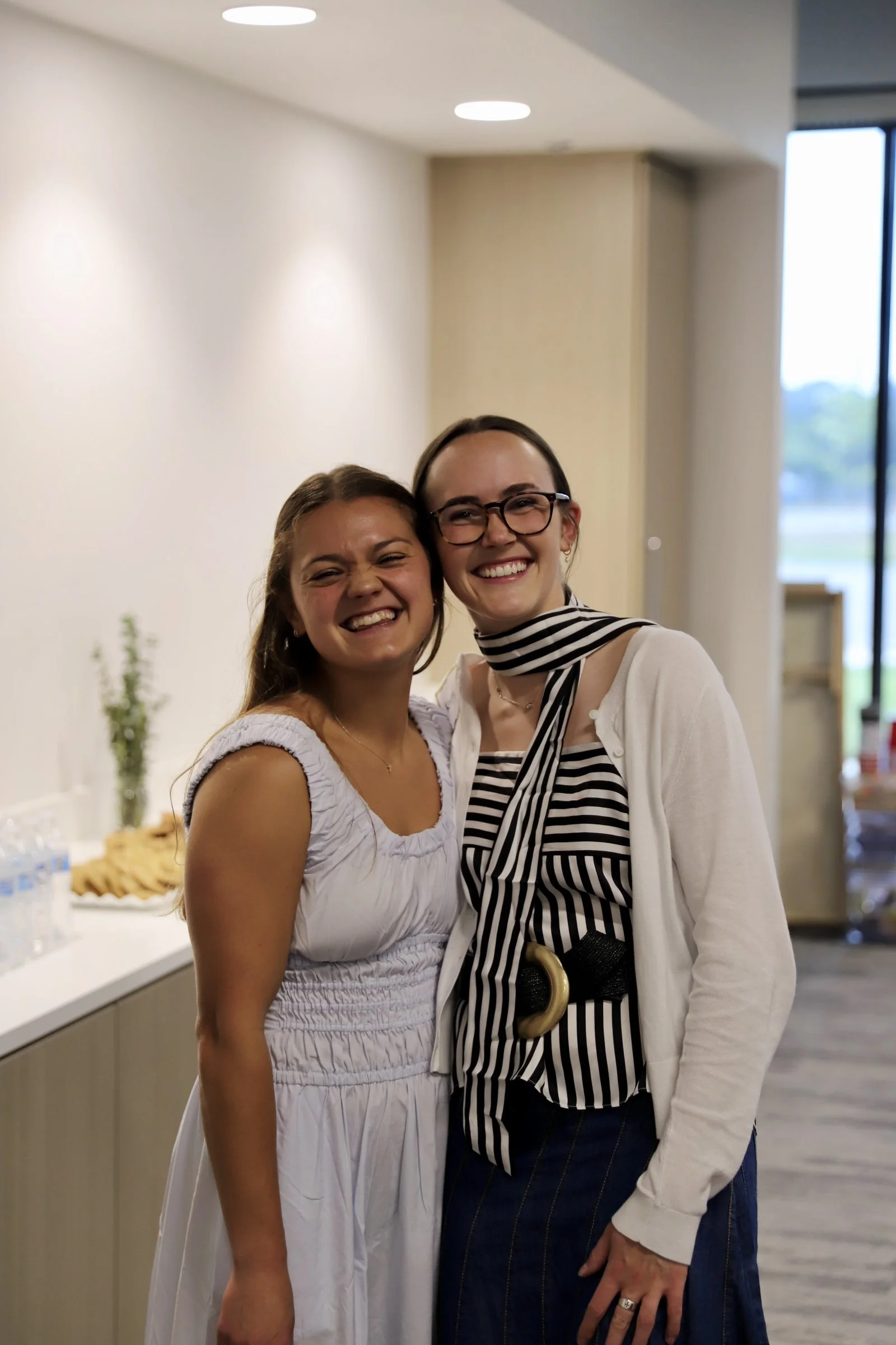 Two women smiling and standing close together indoors near a countertop, with a window and a bright space visible in the background.