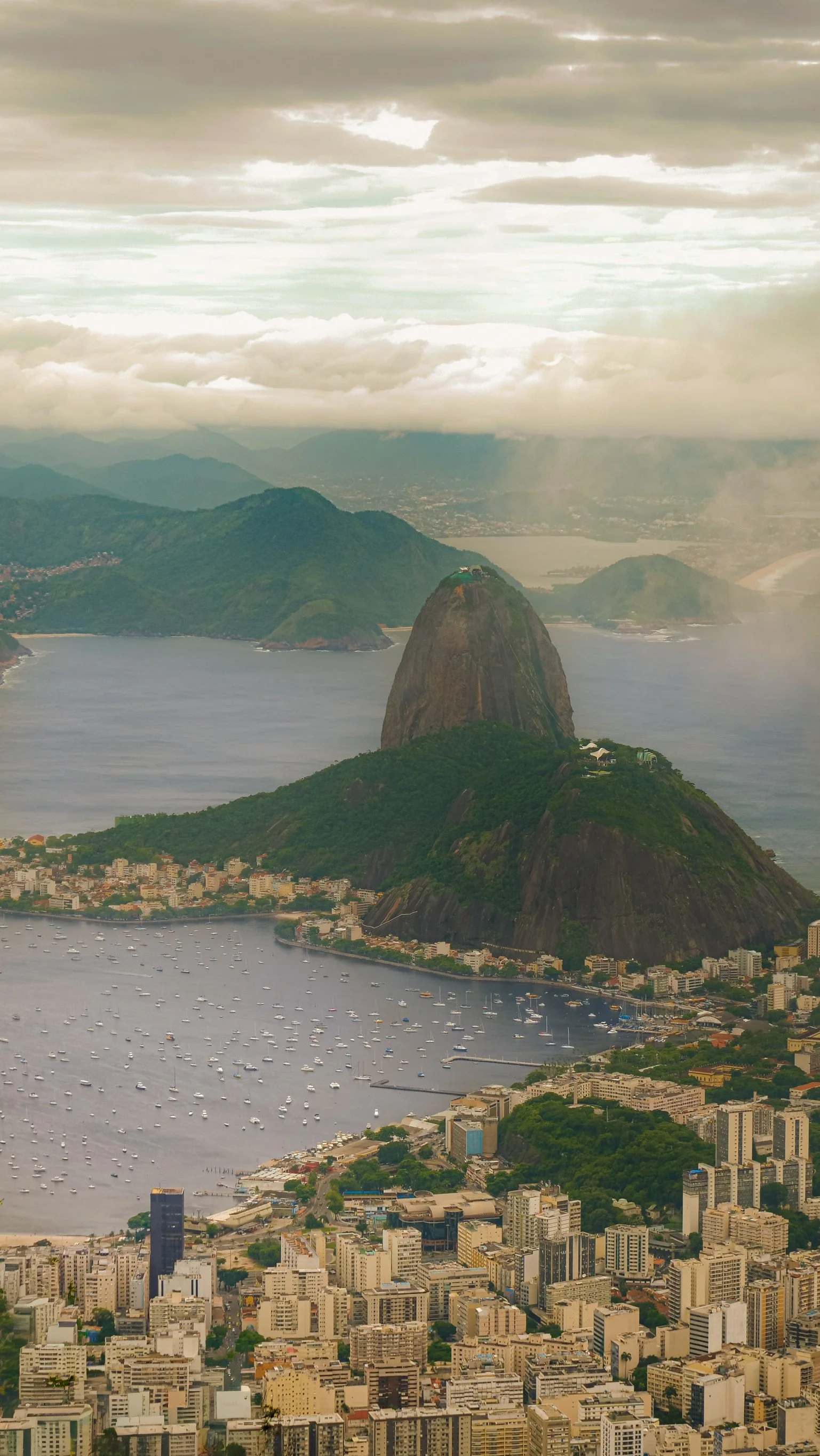 Aerial view of Rio de Janeiro, Brazil, showing Sugarloaf Mountain, city buildings, and a harbor filled with boats under a cloudy sky.