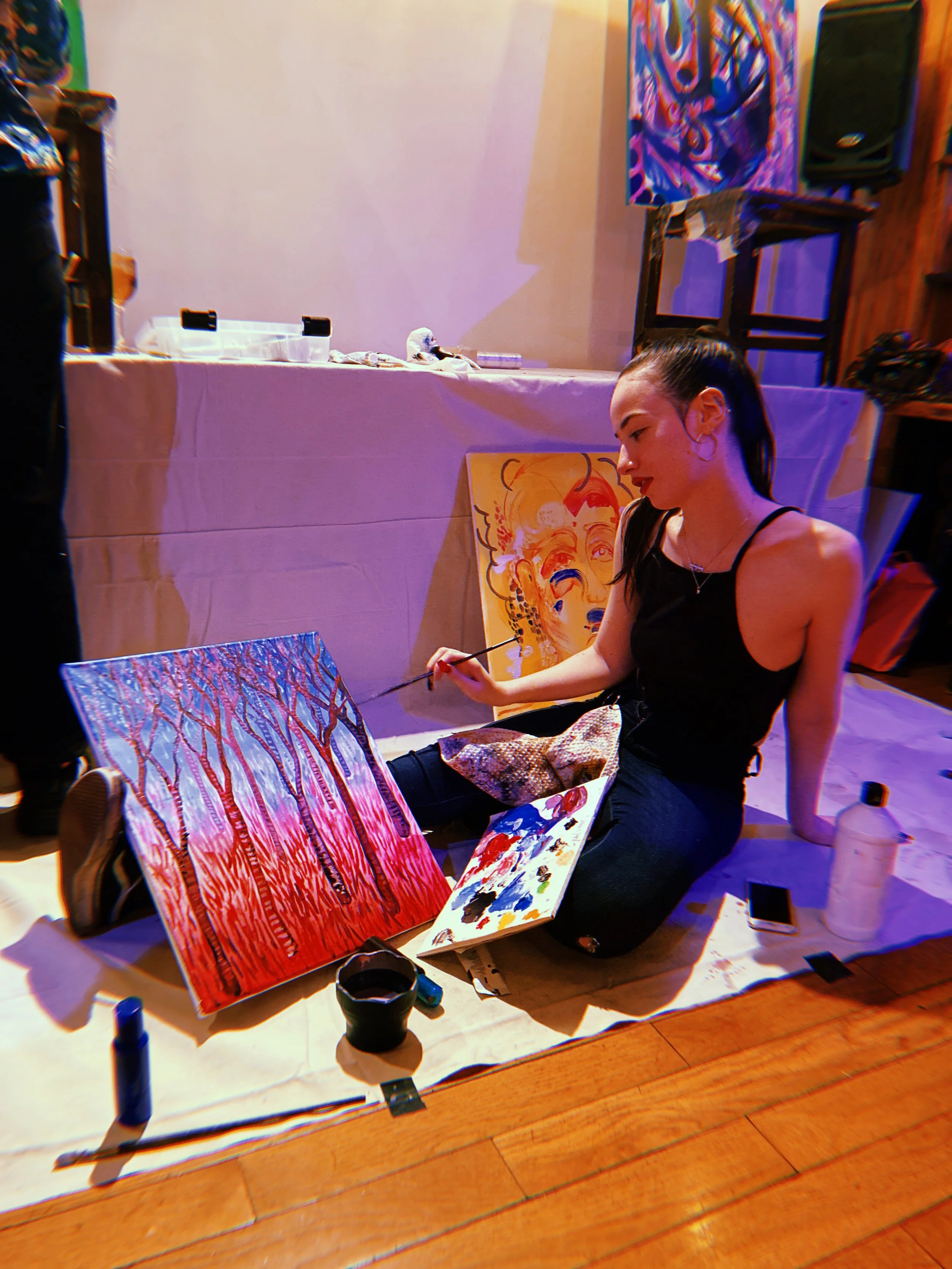 A woman sitting cross-legged on the floor painting on a canvas with a paintbrush. The canvas features a painting with bright red, orange, and purple colors. There are painting supplies, including a palette and bottles, on the paper spread out in front of her. Behind her, various artworks are displayed, and there are chairs and a table in the background.