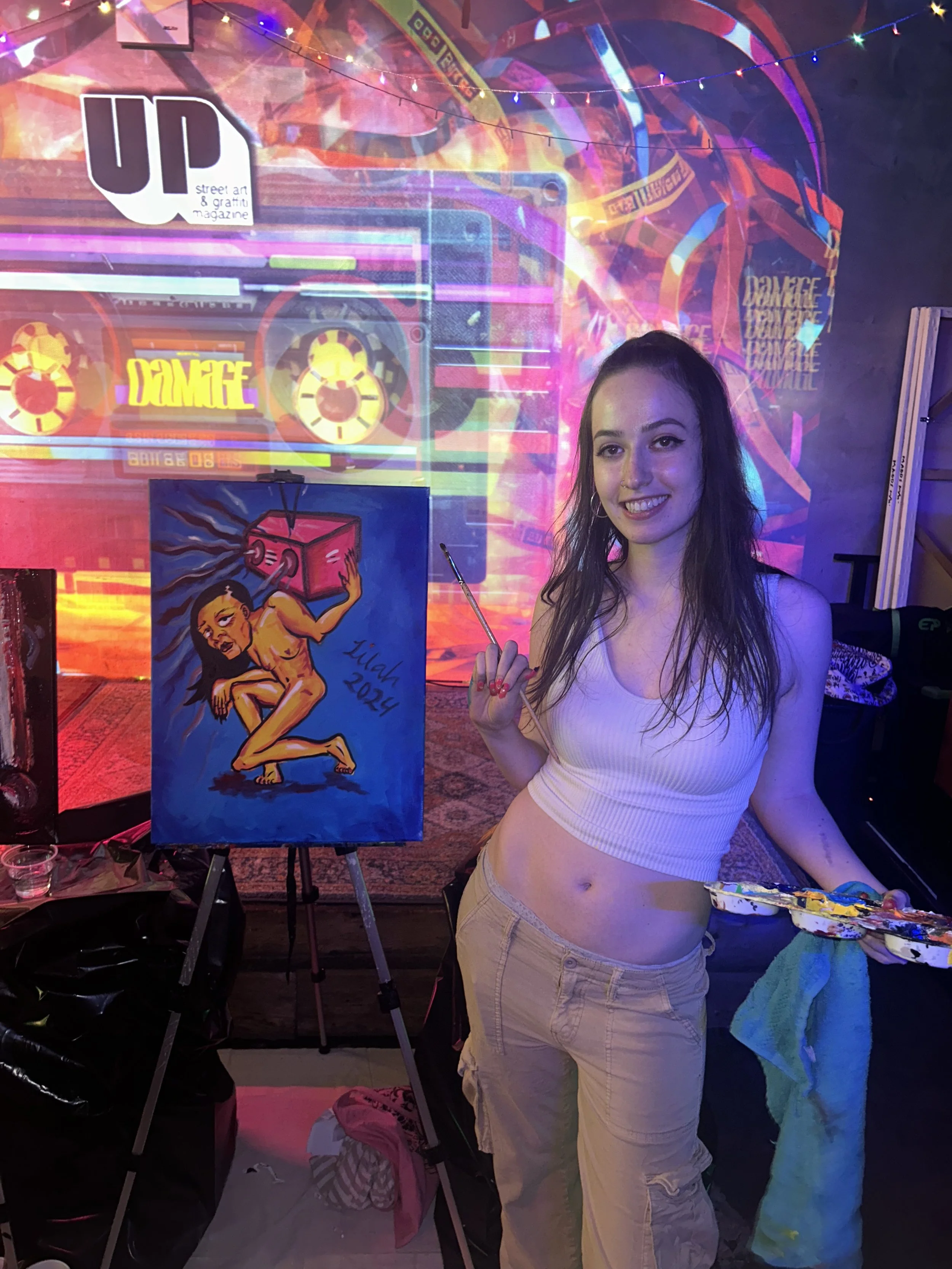 A young woman with long hair and wearing a white tank top and beige cargo pants, holding a paintbrush and smiling, standing in front of a colorful graffiti art piece featuring a naked woman holding a pink box on a blue background, at an art event or studio.