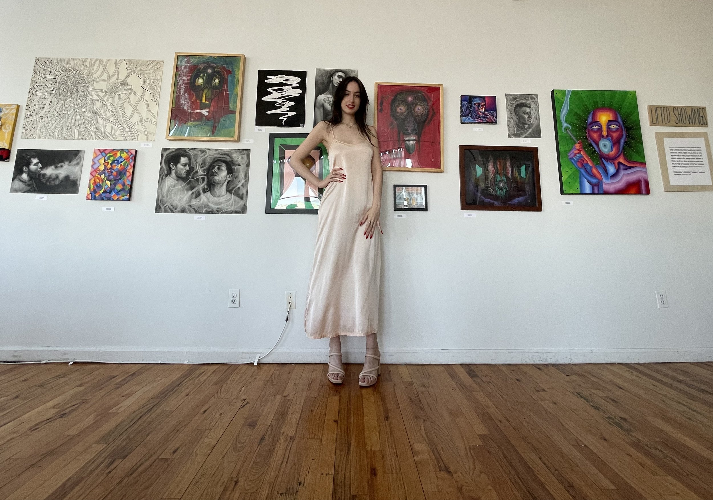 A woman in a satin dress and high heels stands with one hand on her hip in front of a wall with various artworks displayed, including portraits and abstract paintings.