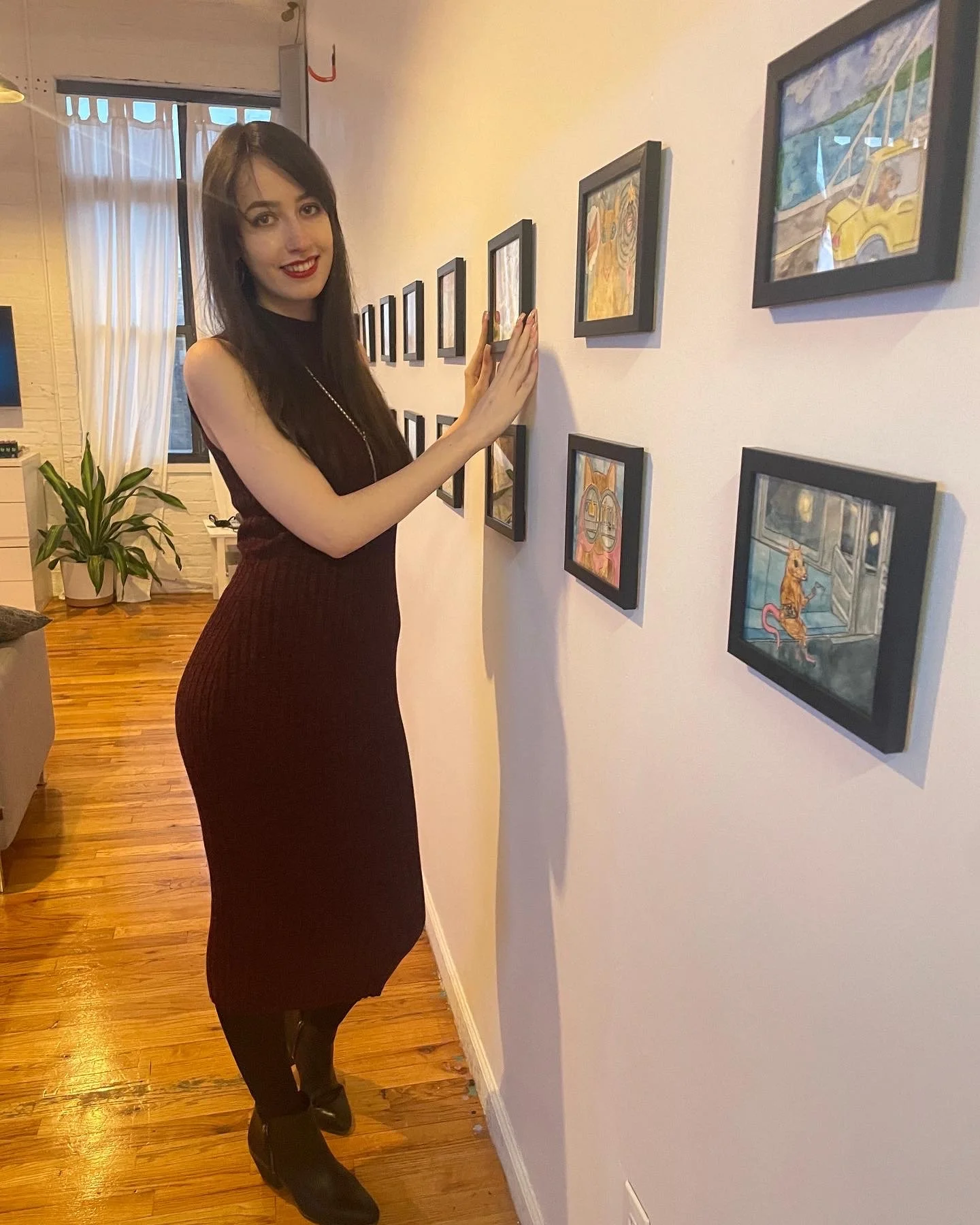 A woman with long dark hair and red lipstick is looking at artwork displayed on a white wall in an art gallery. She is wearing a sleeveless maroon dress and black boots, and is touching one of the framed paintings with her right hand.