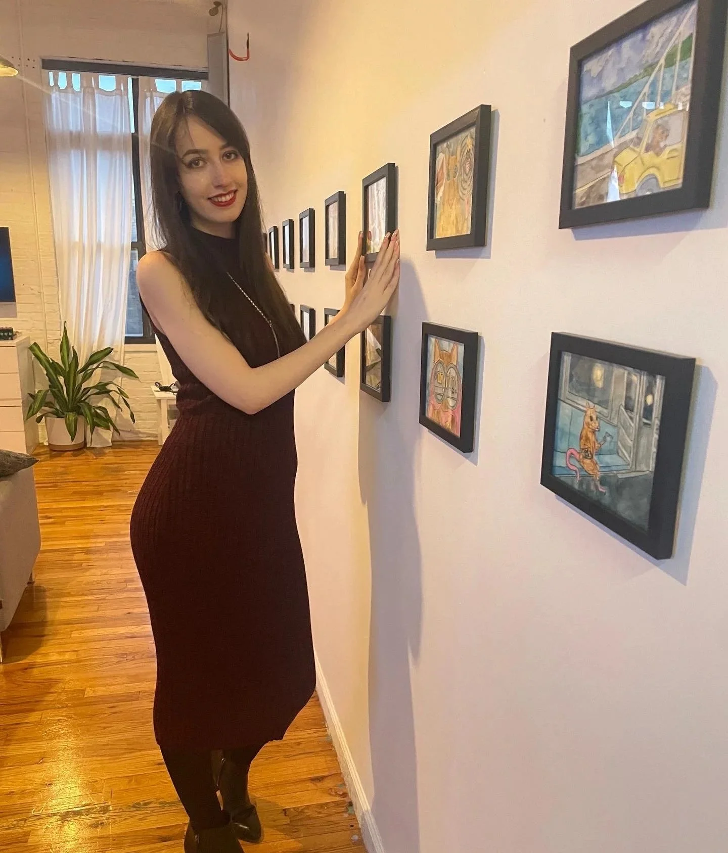 A woman with long dark hair, wearing a sleeveless black top and fitted black skirt, standing in an art gallery, touching a framed painting on the wall, with a smile and red lipstick.