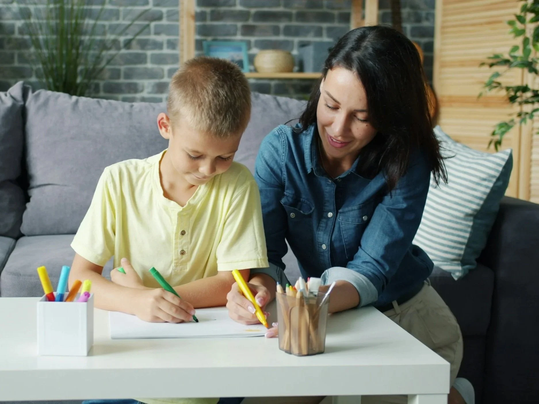 A woman and a young boy sitting at a white table, drawing with colored markers in a modern living room with gray brick walls and greenery.