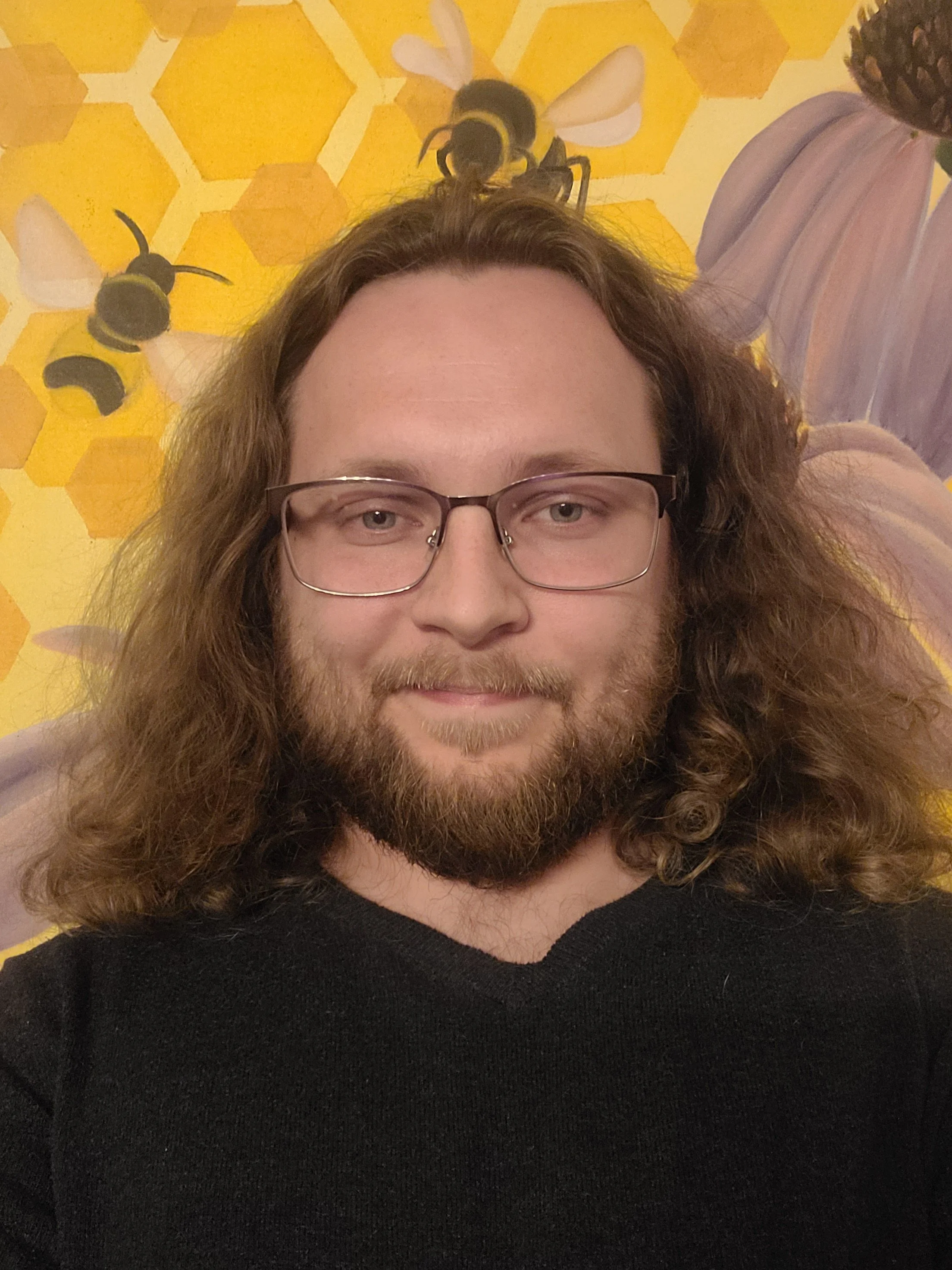 Close-up of a person with long, curly hair and glasses, wearing a black top, with a background of yellow honeycomb pattern and bees, including a bee sculpture or decoration.