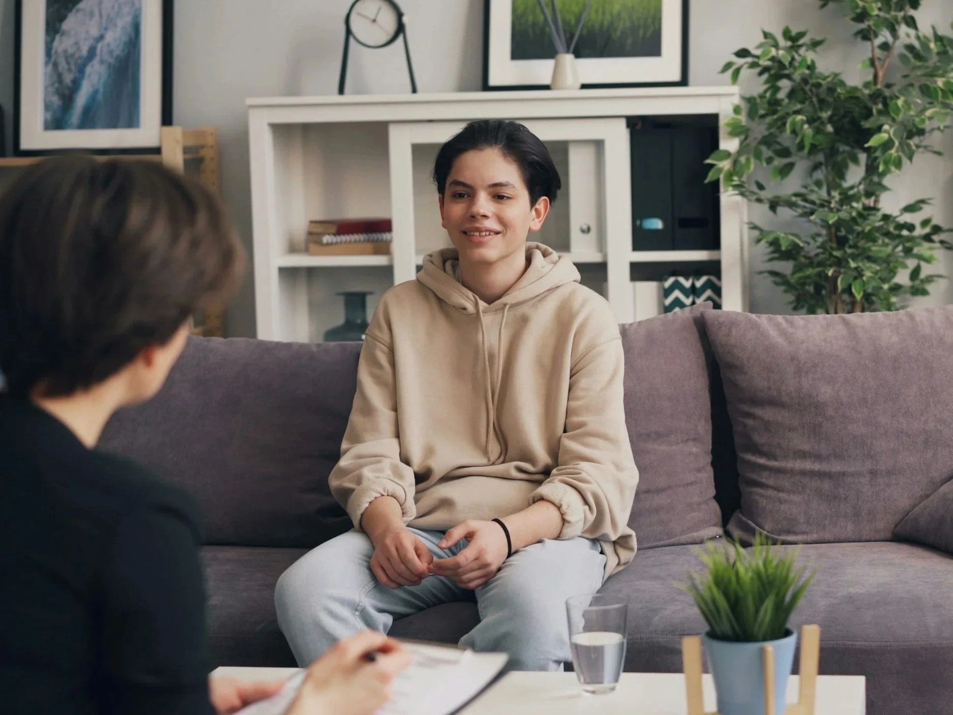 Two young men having a conversation in a living room. One is sitting on a sofa, smiling, wearing a beige hoodie and light-colored pants. The other is partially visible, holding a clipboard and pen, taking notes. The room has framed artwork, a clock, and a large potted plant in the background. There are glasses of water and a small potted plant on the table in front.