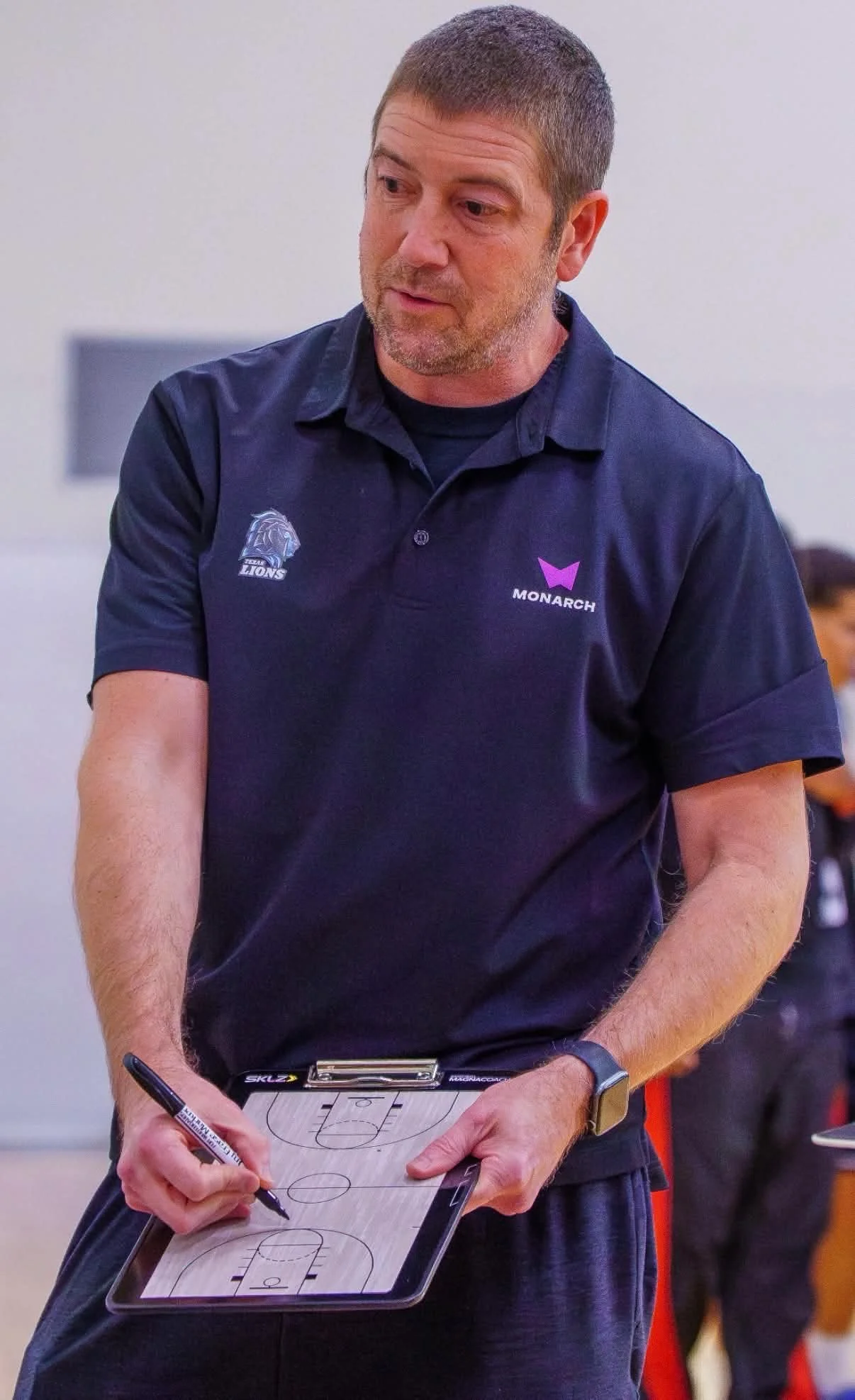 A man in a navy blue polo shirt with a logo on the left chest labeled "LIONS" and a smaller logo on the right labeled "MONARCH" is standing and writing on a small basketball coach’s clipboard with a black marker. The man is in an indoor sports setting and appears to be coaching or instructing.