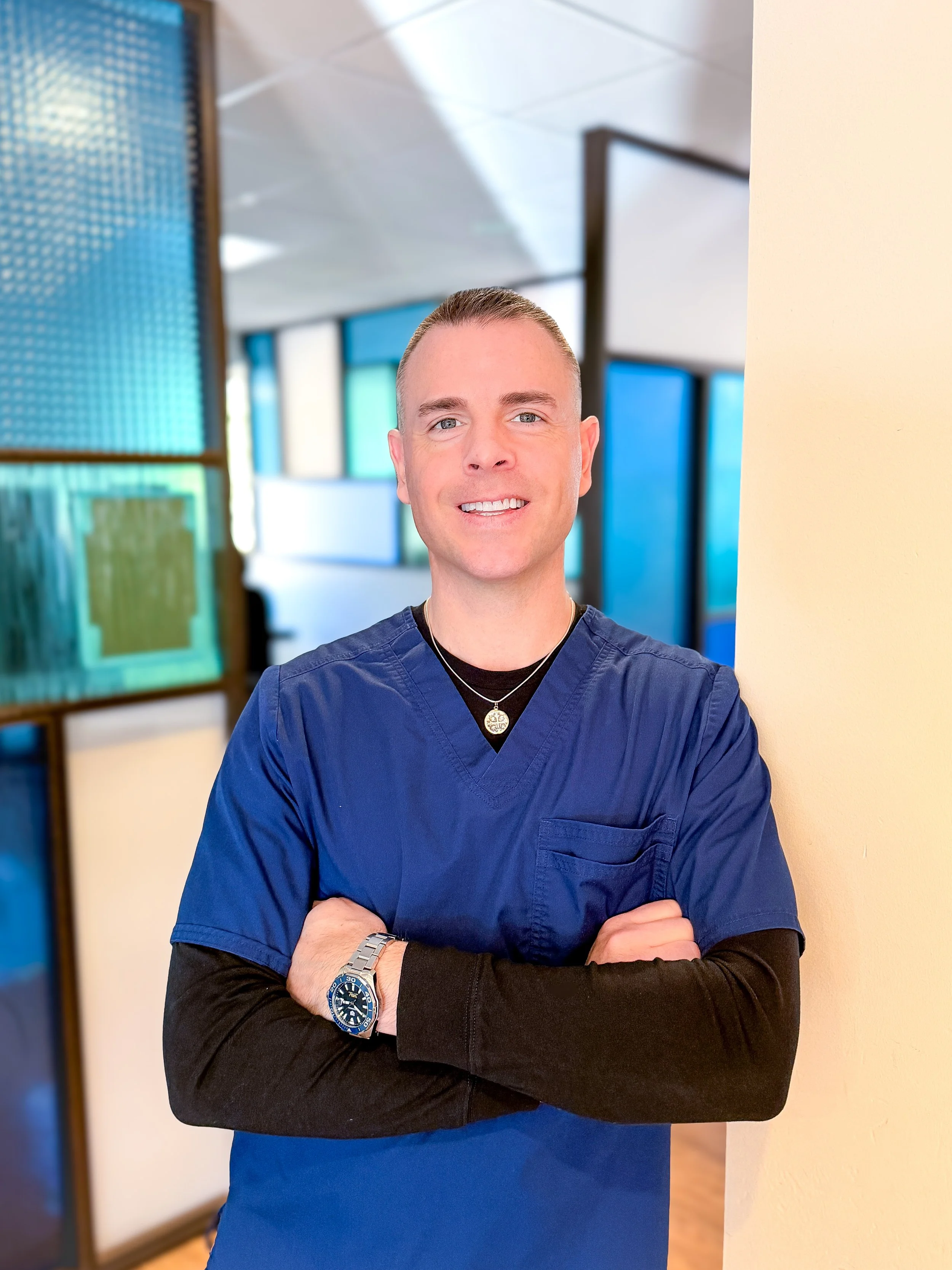 A smiling man in medical scrubs with arms crossed, standing in a colorful, modern healthcare or hospital hallway.