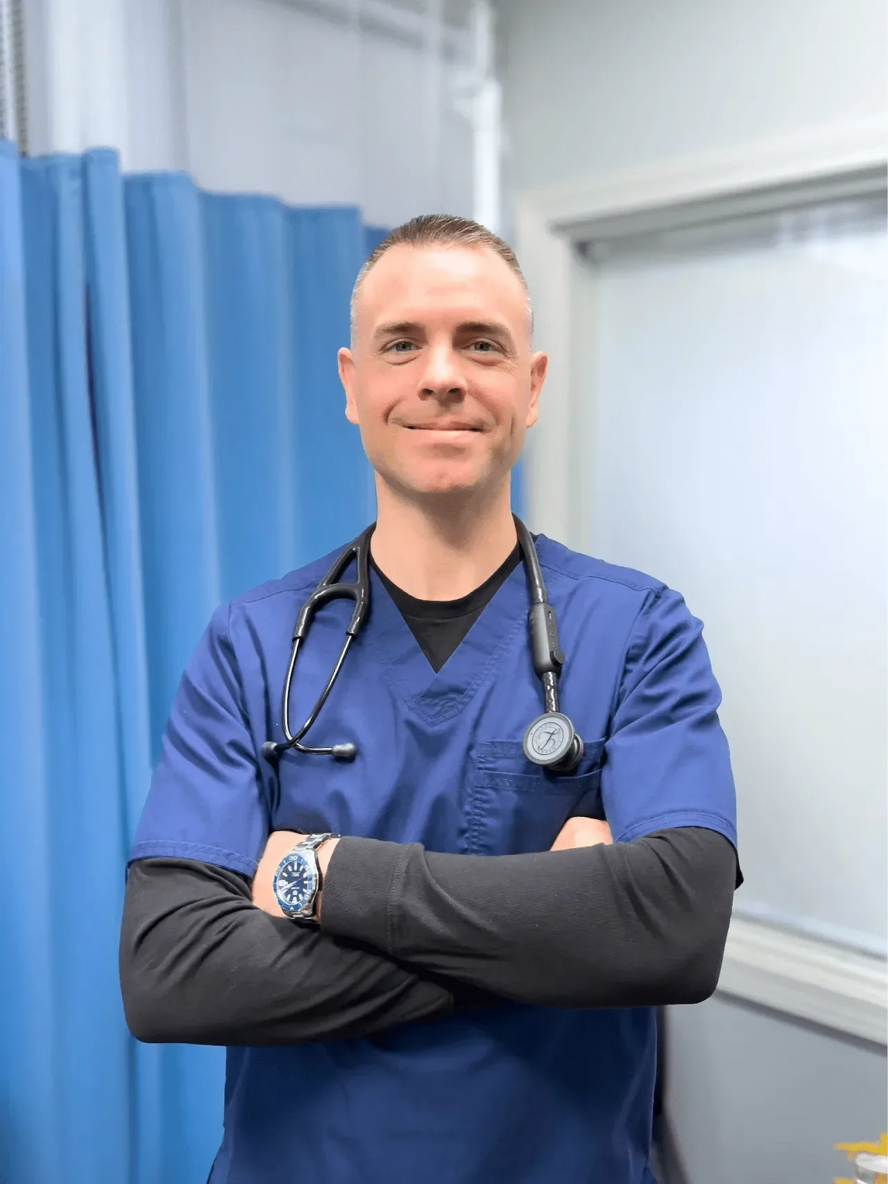 A male healthcare professional in blue scrubs with a stethoscope around his neck, standing with arms crossed in a hospital or medical facility.