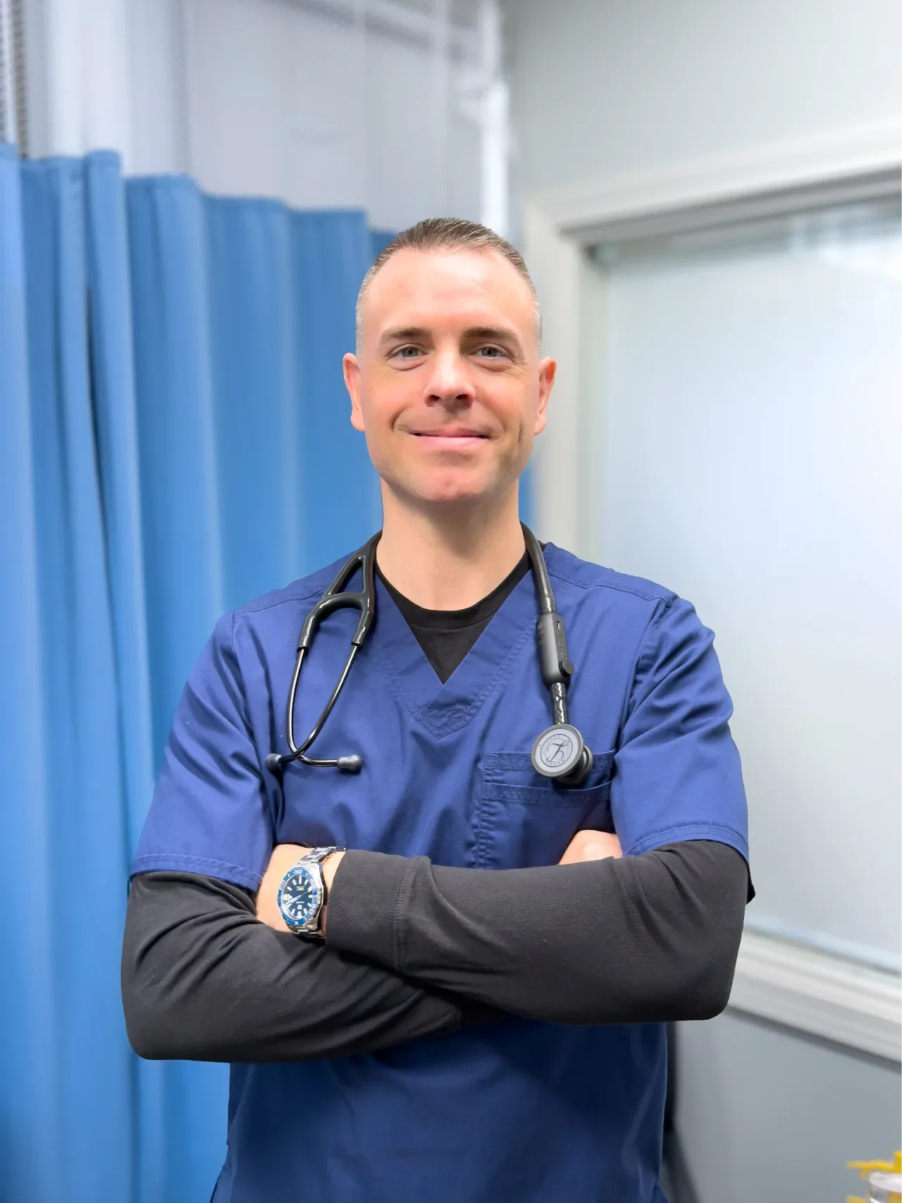 A male healthcare professional with a stethoscope around his neck, wearing blue scrubs and a watch, standing with his arms crossed in a clinical setting with blue curtains and windows in the background.