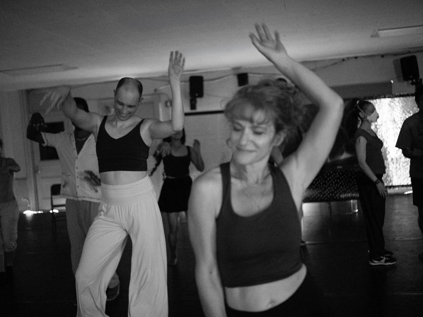 Black-and-white photo of people dancing in a fitness or dance class, with two women raising their arms in the foreground.