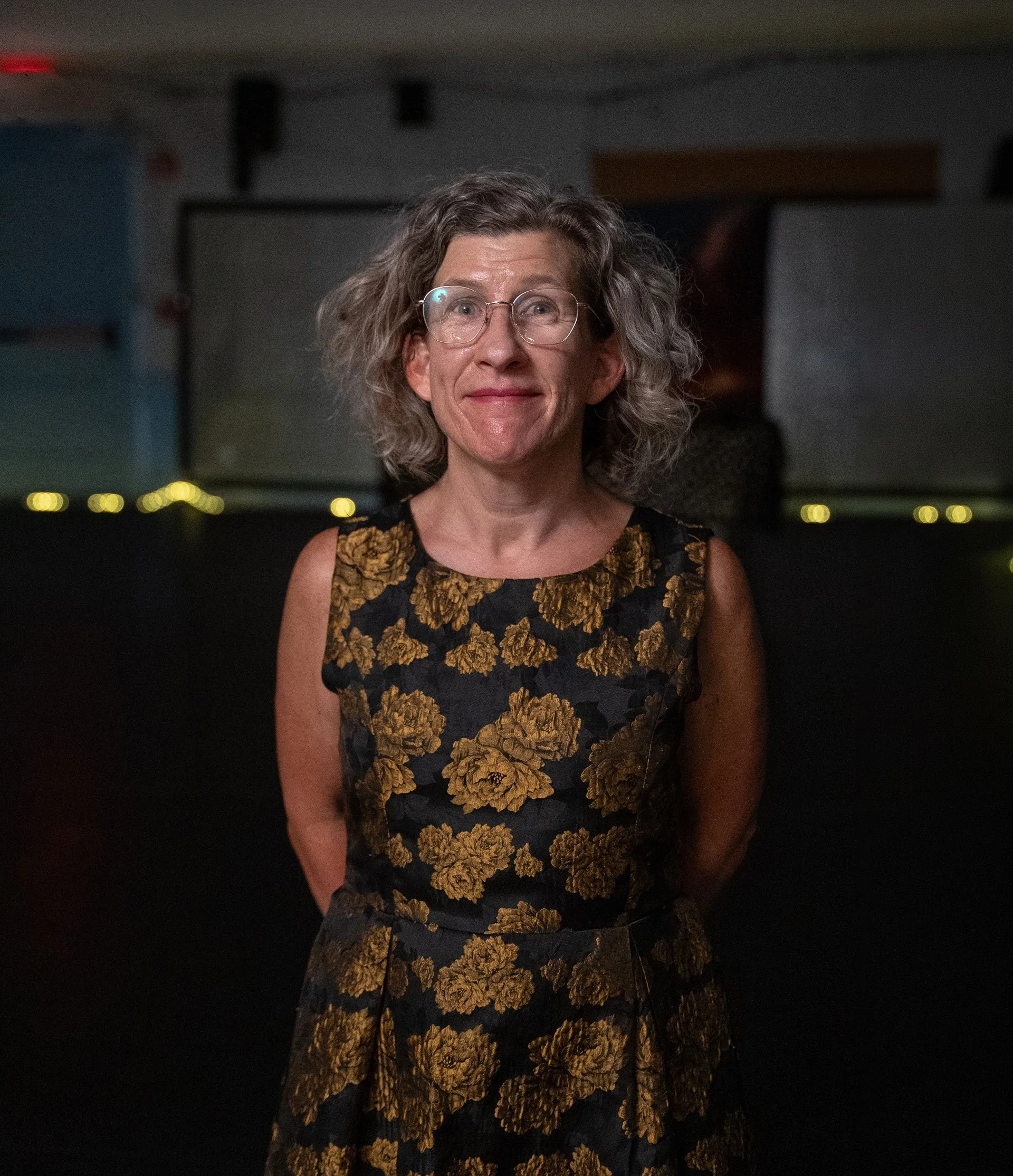 Meghan smiling with glasses and wearing a black dress with gold floral patterns indoors