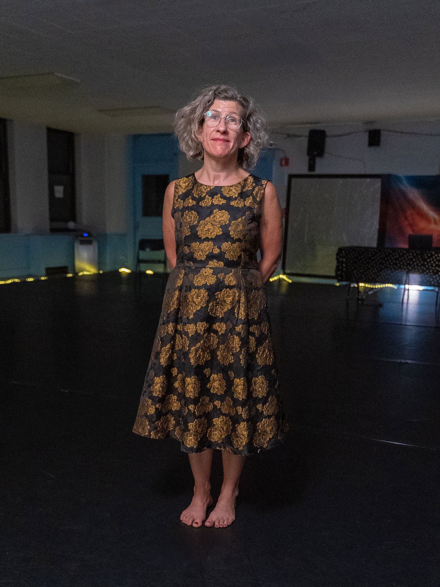 Meghan wearing glasses and a black-and-gold floral dress standing barefoot in a dimly lit gallery or event space