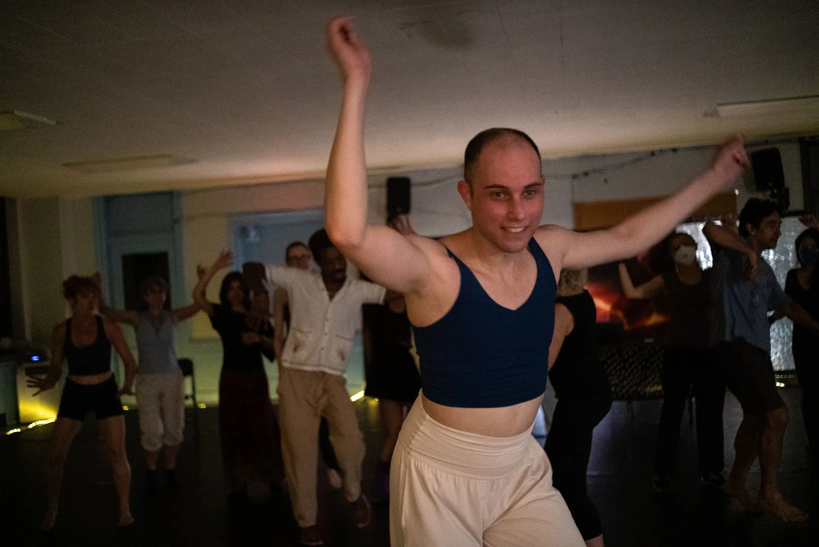 People dancing at an indoor party, with a smiling dancer in a navy tank top and white pants in the foreground.