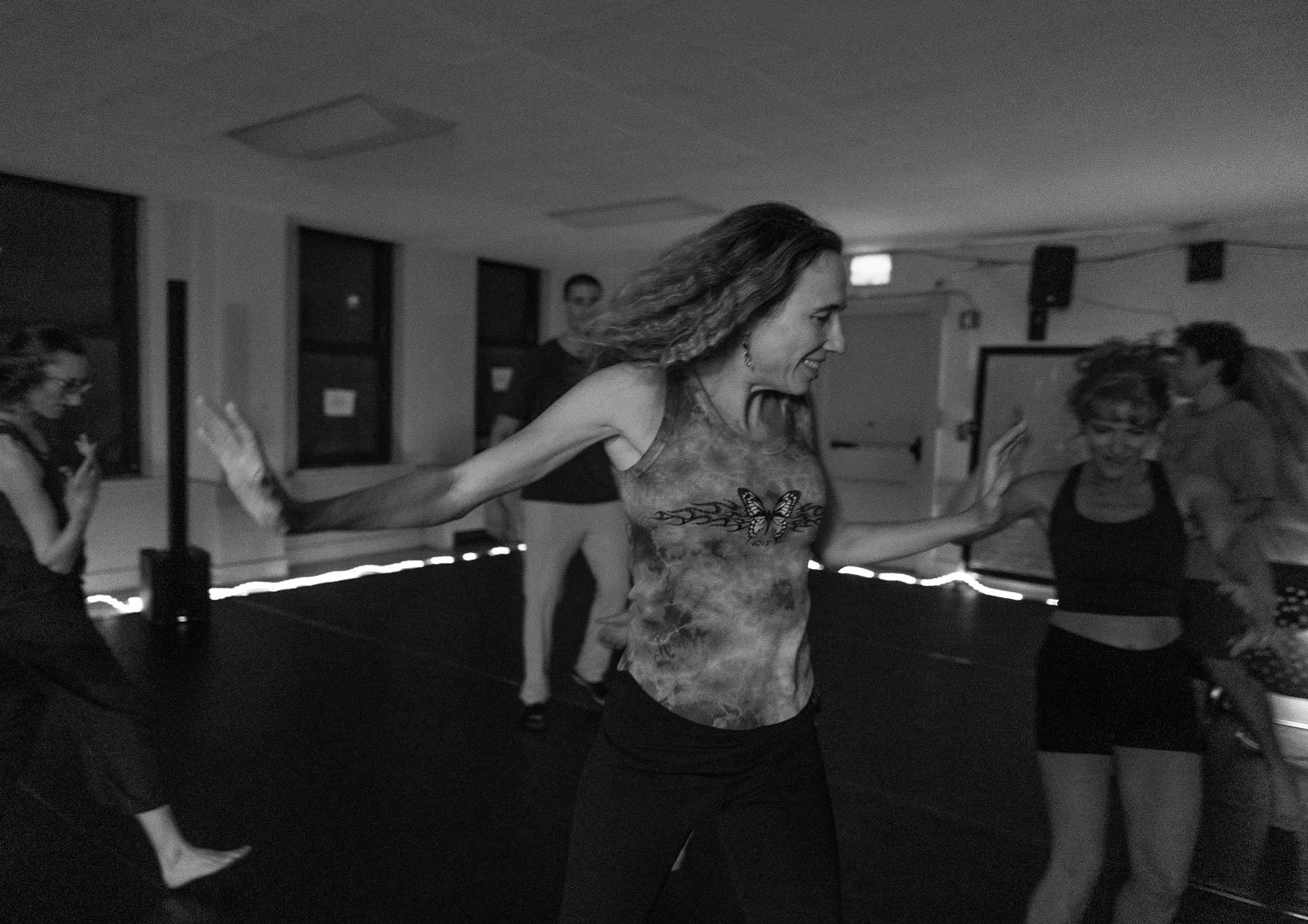 People dancing in a dimly lit studio, led by a woman with outstretched arms.