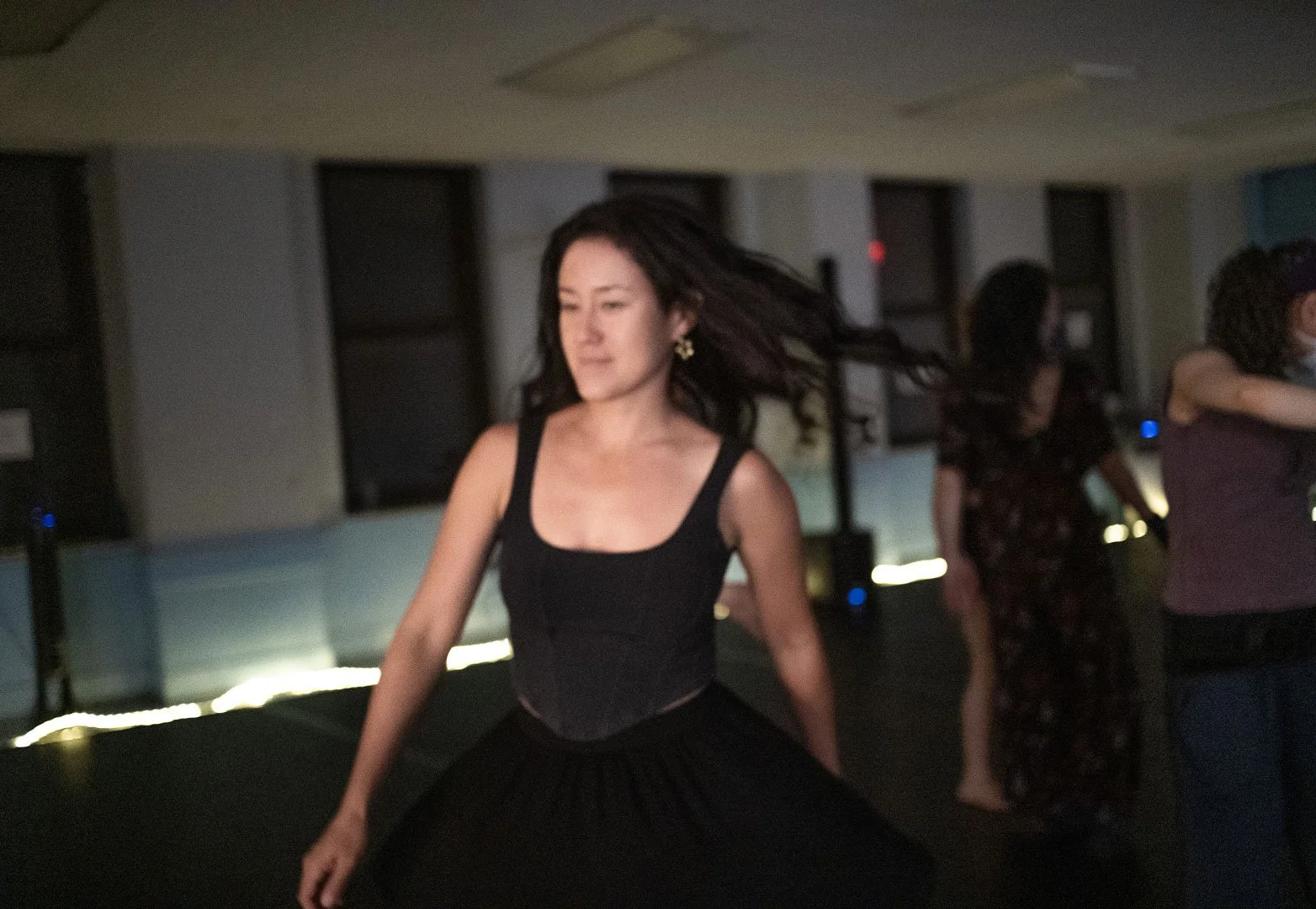 Dancer in a black dress performing in a dimly lit indoor space with other dancers in the background