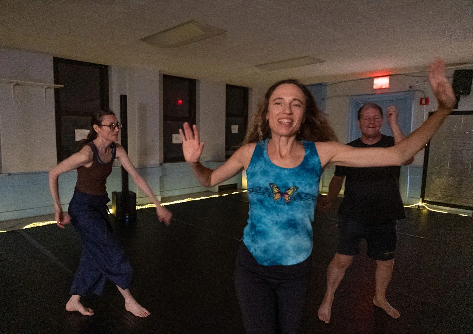 Three barefoot adults dancing in a dimly lit studio, with a smiling woman in a blue butterfly tank top in the foreground.