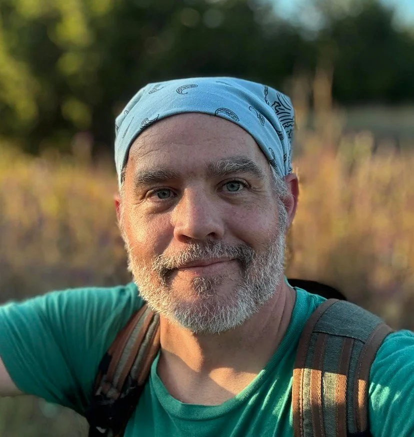 A middle-aged man with a beard and mustache taking a selfie outdoors. He's wearing a light blue bandana and a teal shirt, with a backpack on his shoulders. The background features trees and a sunny, natural setting.