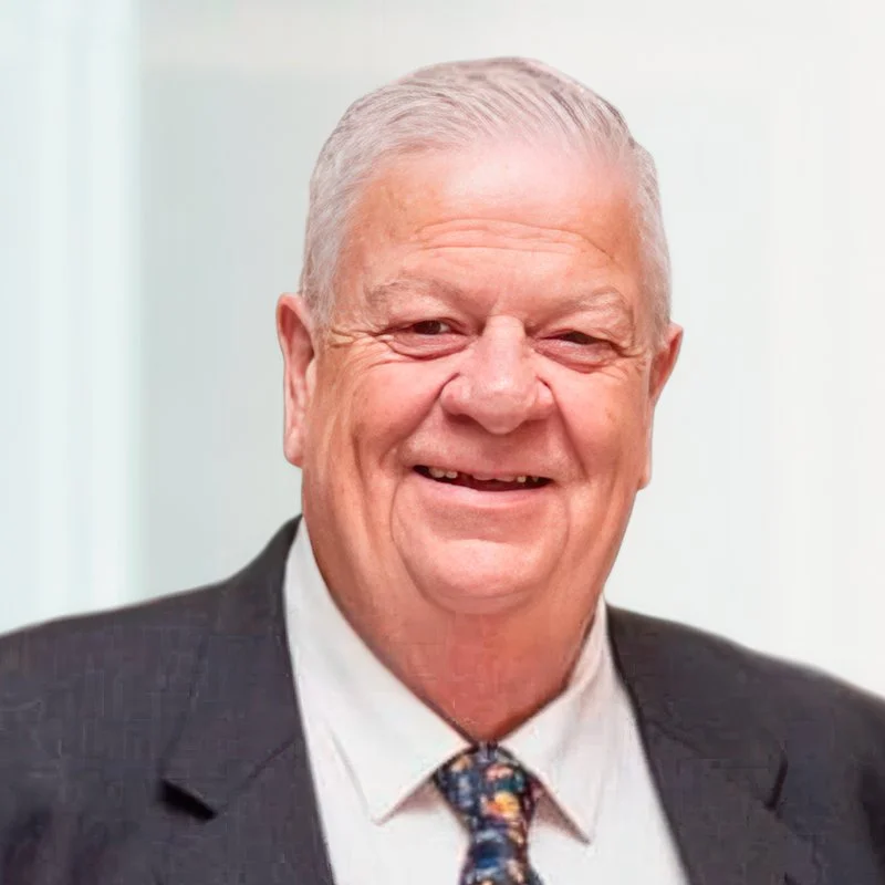 A smiling elderly man with white hair in a suit and tie, standing against a neutral background.