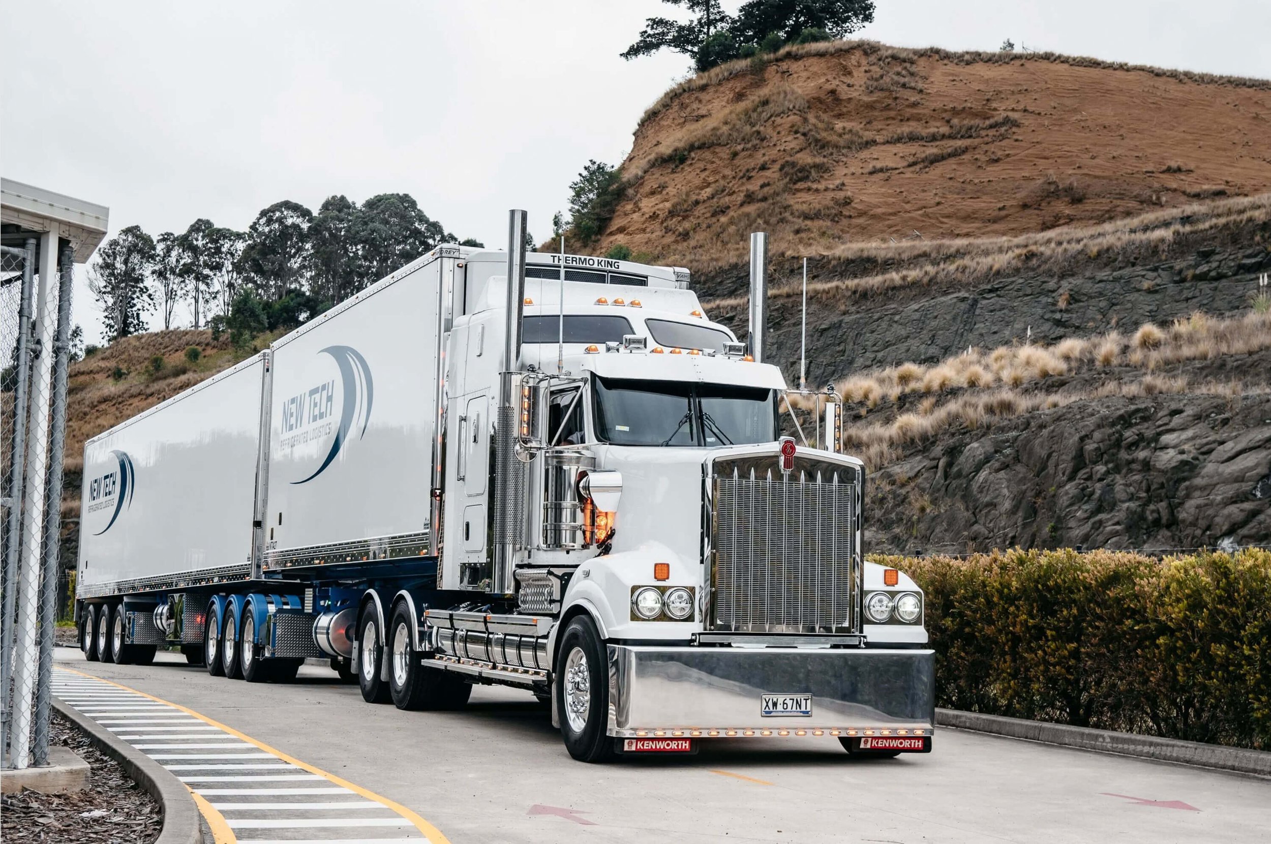A large white semi-truck with long trailer driving on a road, surrounded by a rocky hillside and trees, overcast sky.