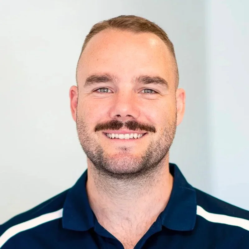 A young man with short hair, a mustache, and a beard, smiling at the camera, wearing a navy blue shirt with white stripes on the shoulders, standing against a plain white background.