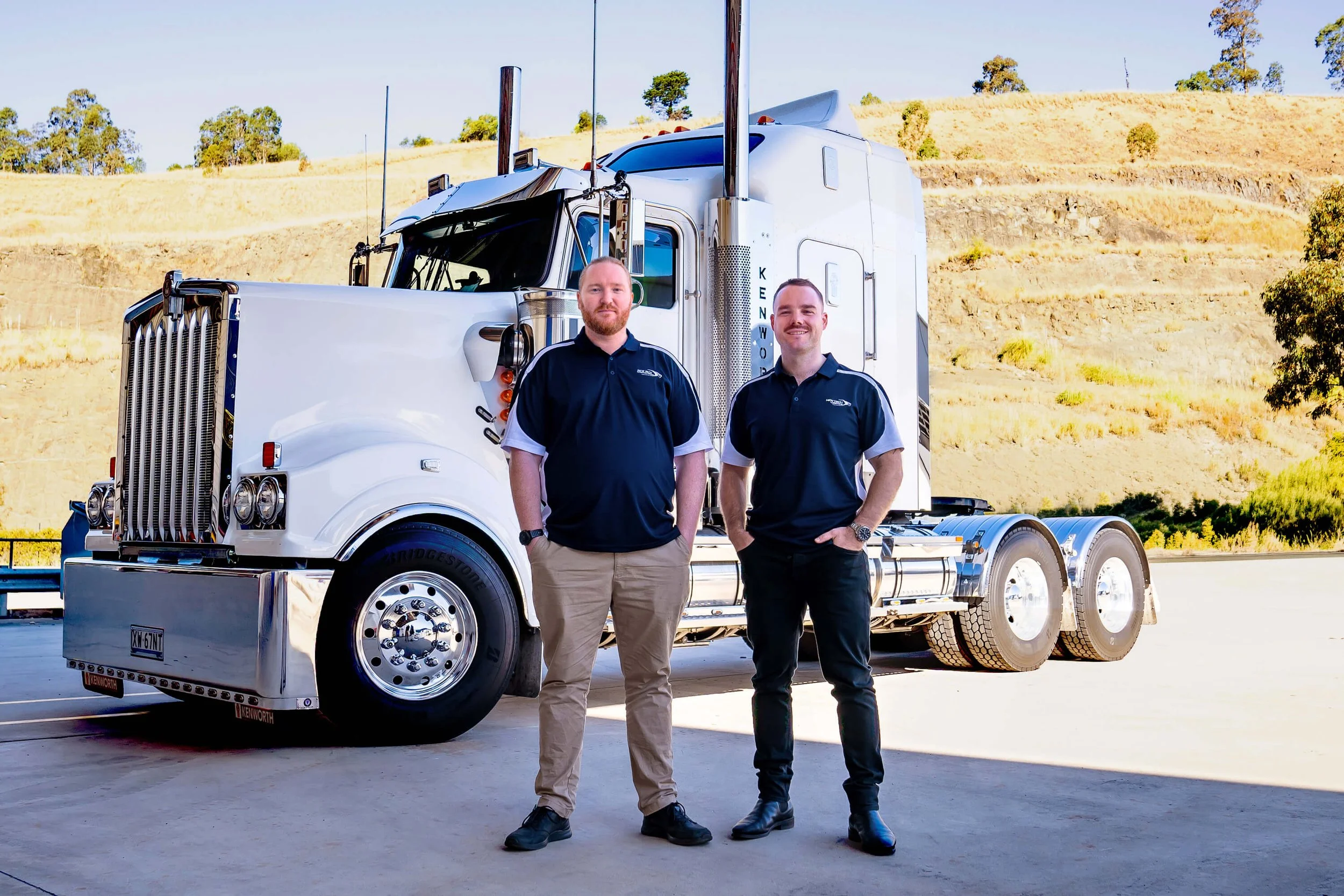 Two men standing in front of a large white semi-truck truck outdoors in a sunny, hilly landscape. They are smiling and wearing black polo shirts with a company logo, and khaki and black pants respectively.